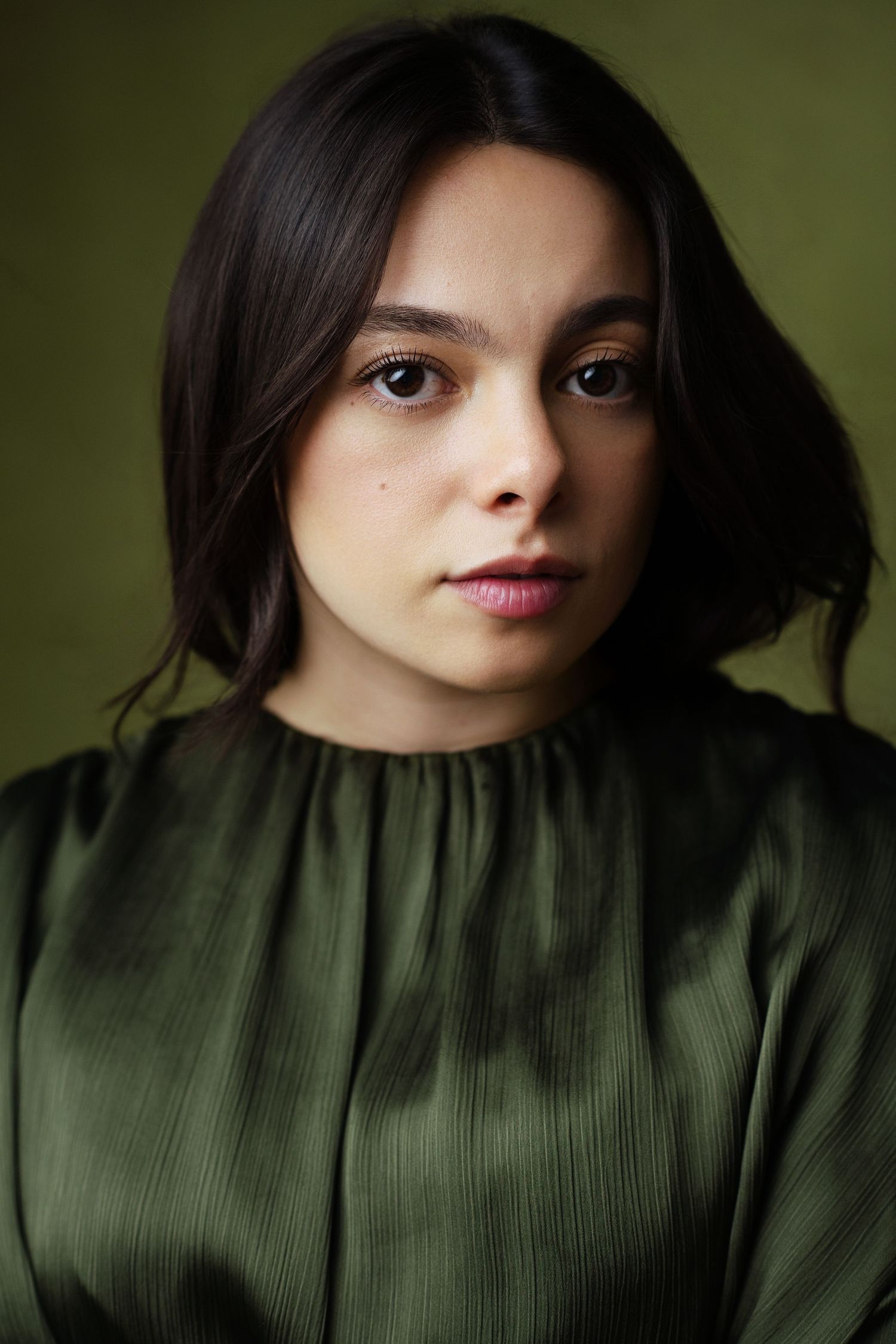 Young woman with dark hair wearing a green satin blouse poses against an olive green background.
