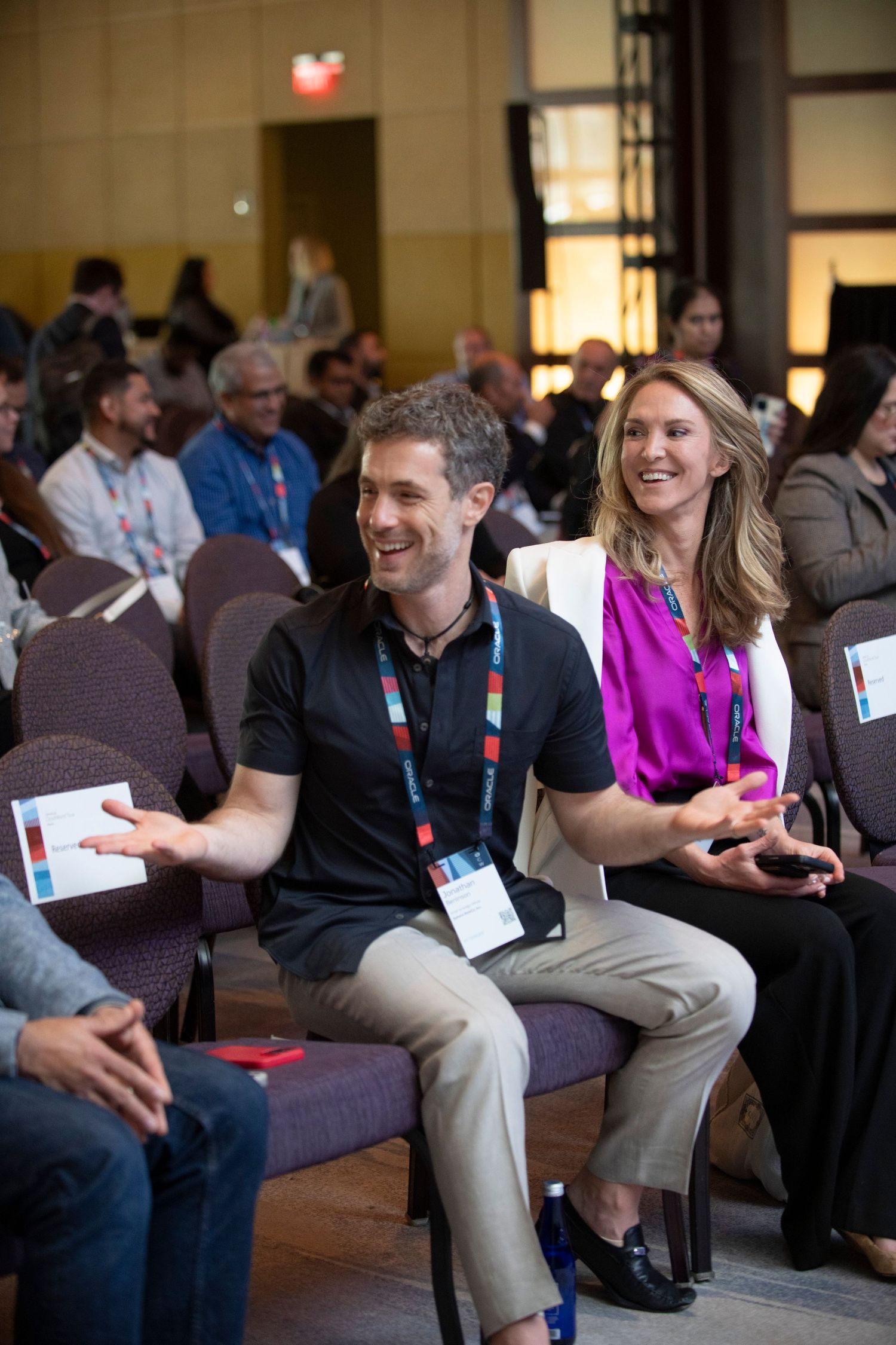 Two professionals laughing and engaged at a conference, wearing lanyards, seated among a crowd of attendees in a meeting hall.