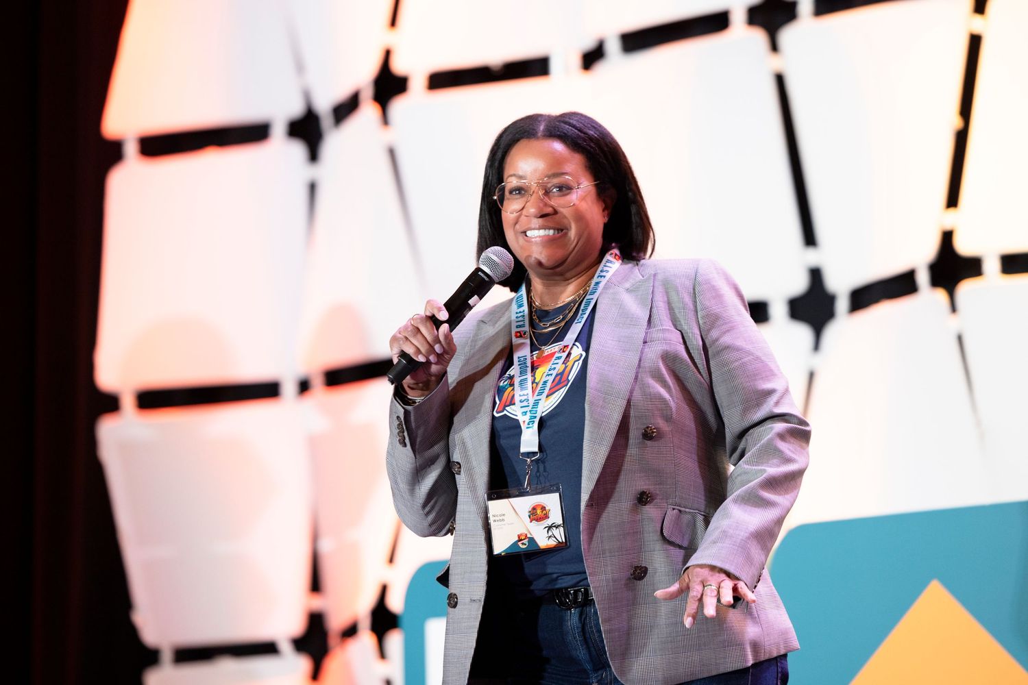 Woman in lavender blazer speaks into microphone on stage at a professional conference event.