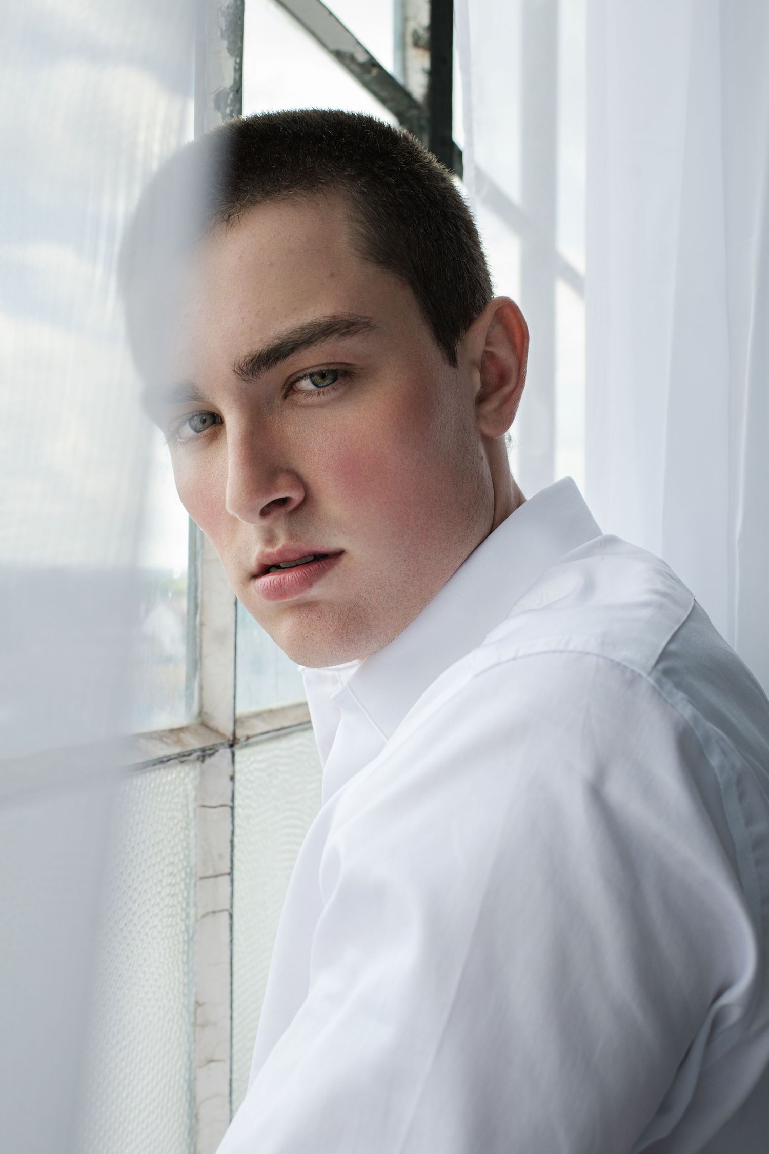 Young man with short hair in white shirt leaning against window with sheer curtains in soft natural light.
