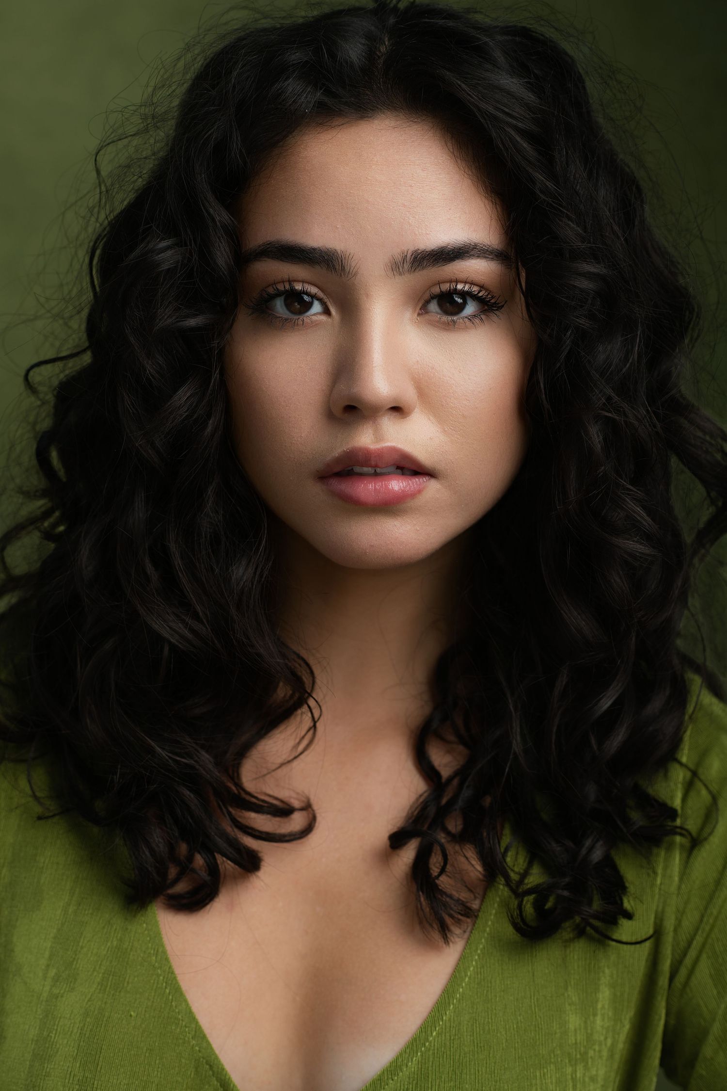 Young woman with curly black hair and green eyes wearing a green top, posing against a dark green background.