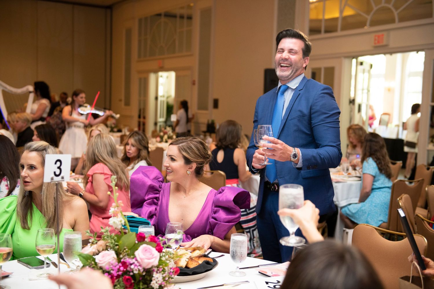 Guests laughing and socializing at an elegant wedding reception banquet hall with floral centerpieces and table number cards.