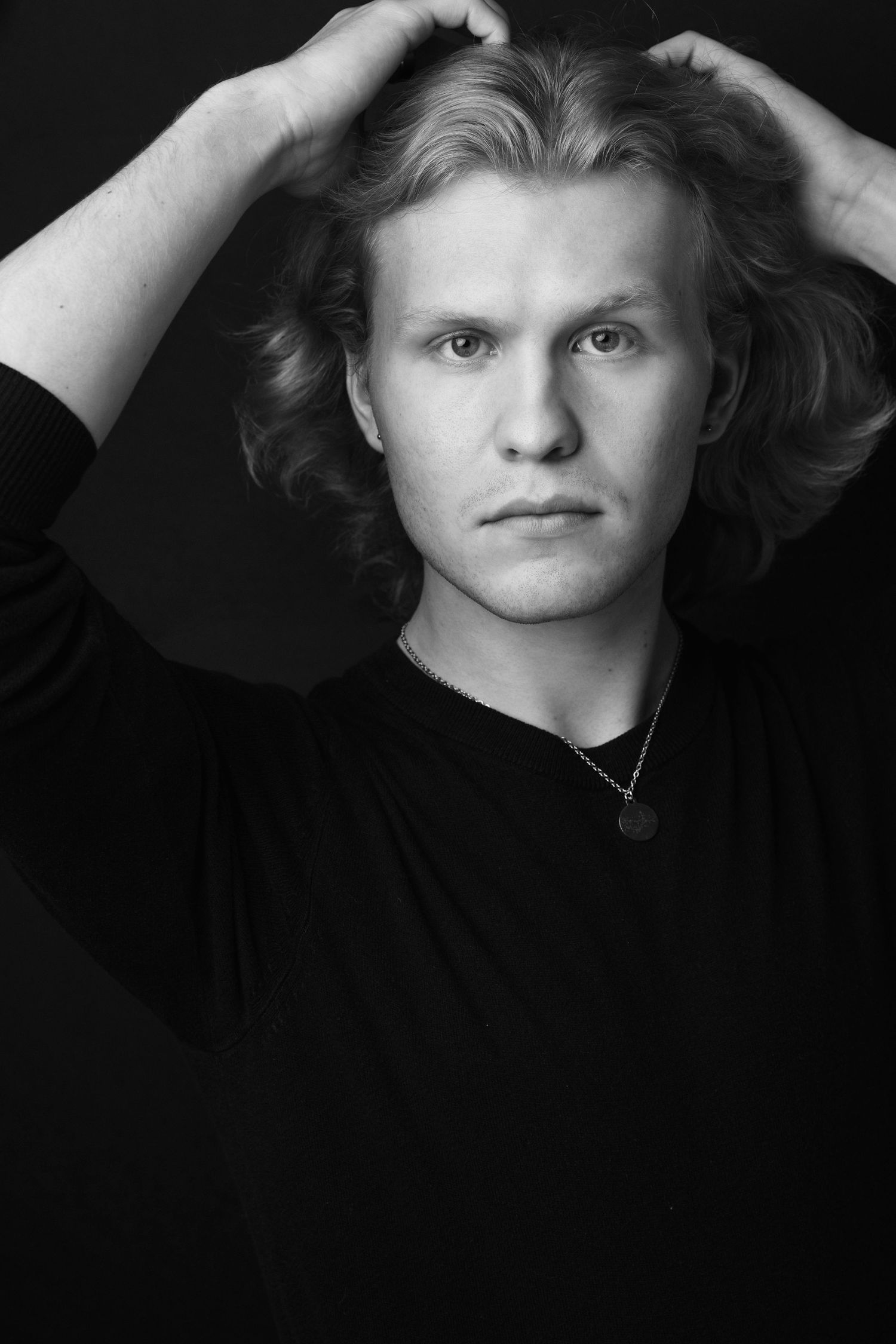 Black and white portrait of a young man with wavy blonde hair, hands raised to his head, wearing a black sweater.