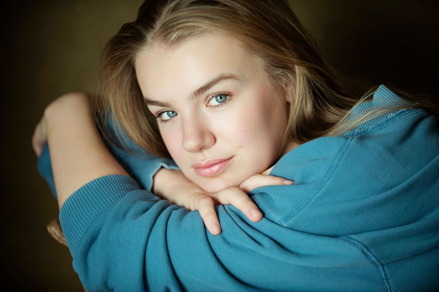 Young blonde woman with green eyes resting chin on hands wearing a blue sweatshirt.