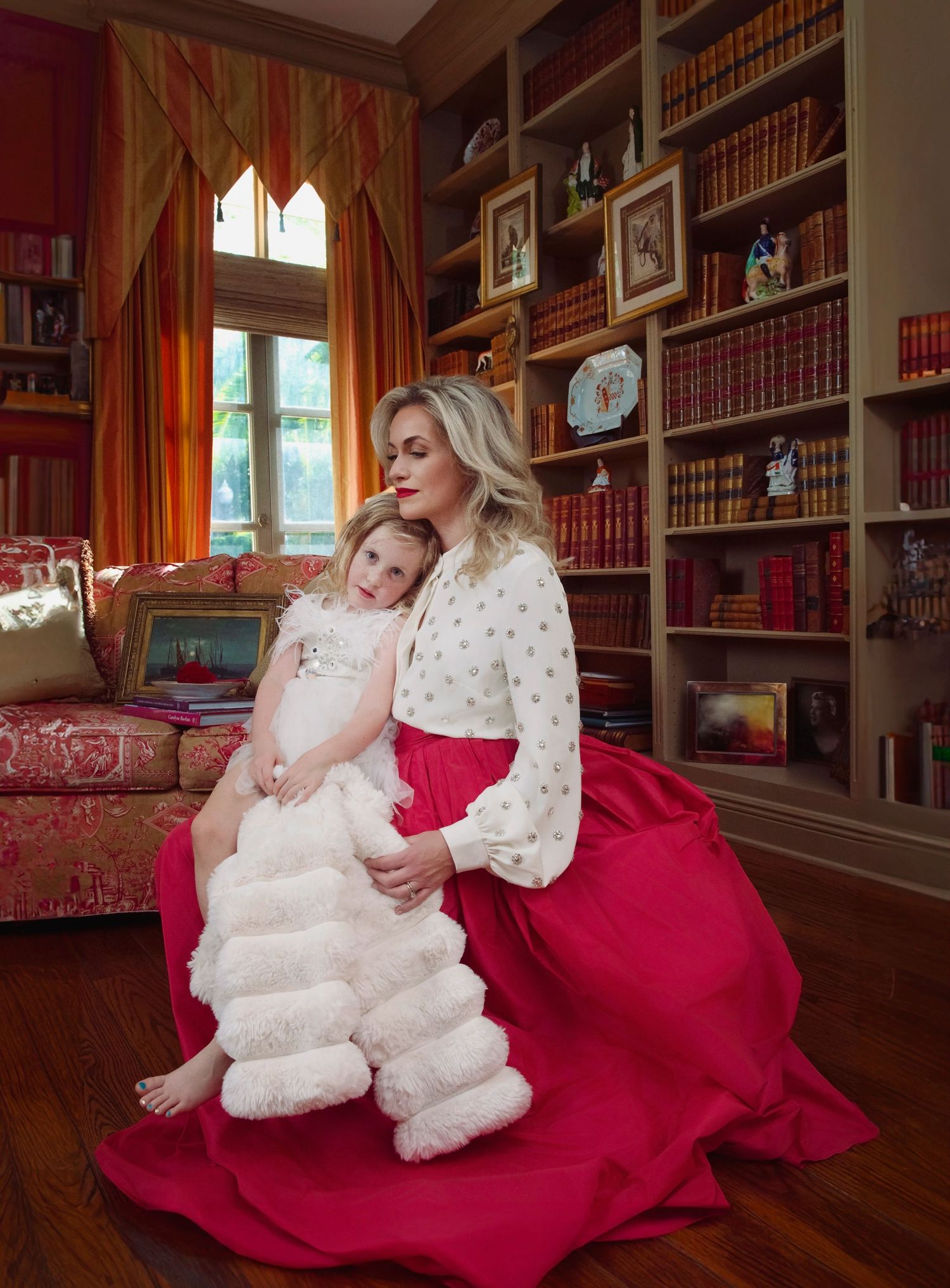 Woman in red skirt and white top holds young girl in white dress in cozy library room.