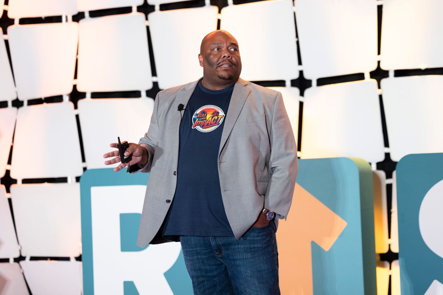 Man in blazer and graphic tee speaks on stage at a colorful event backdrop.