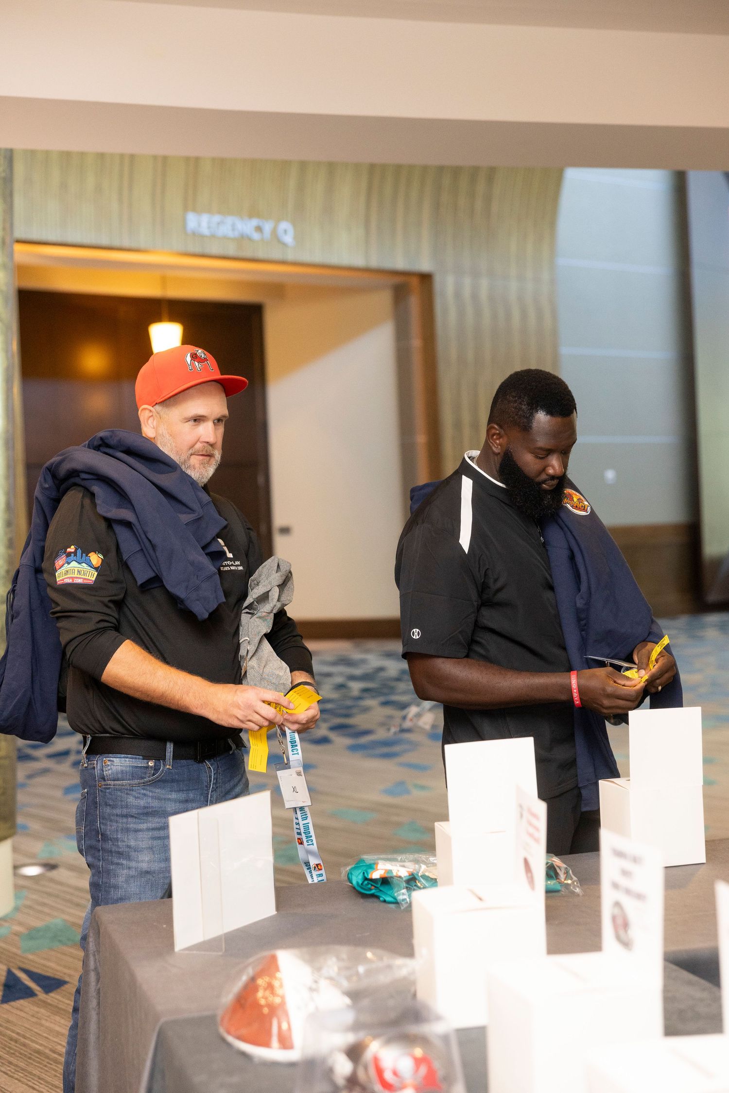 Two men in black jackets examine items at a registration table inside a hotel conference room.