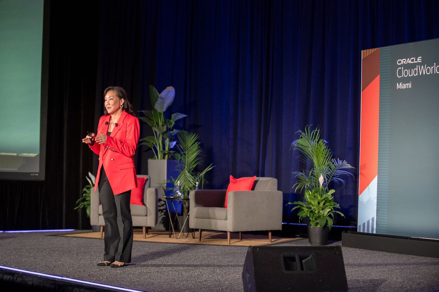 A woman in a red blazer presents on stage at Oracle CloudWorld Miami conference with seating and tropical plants behind her.