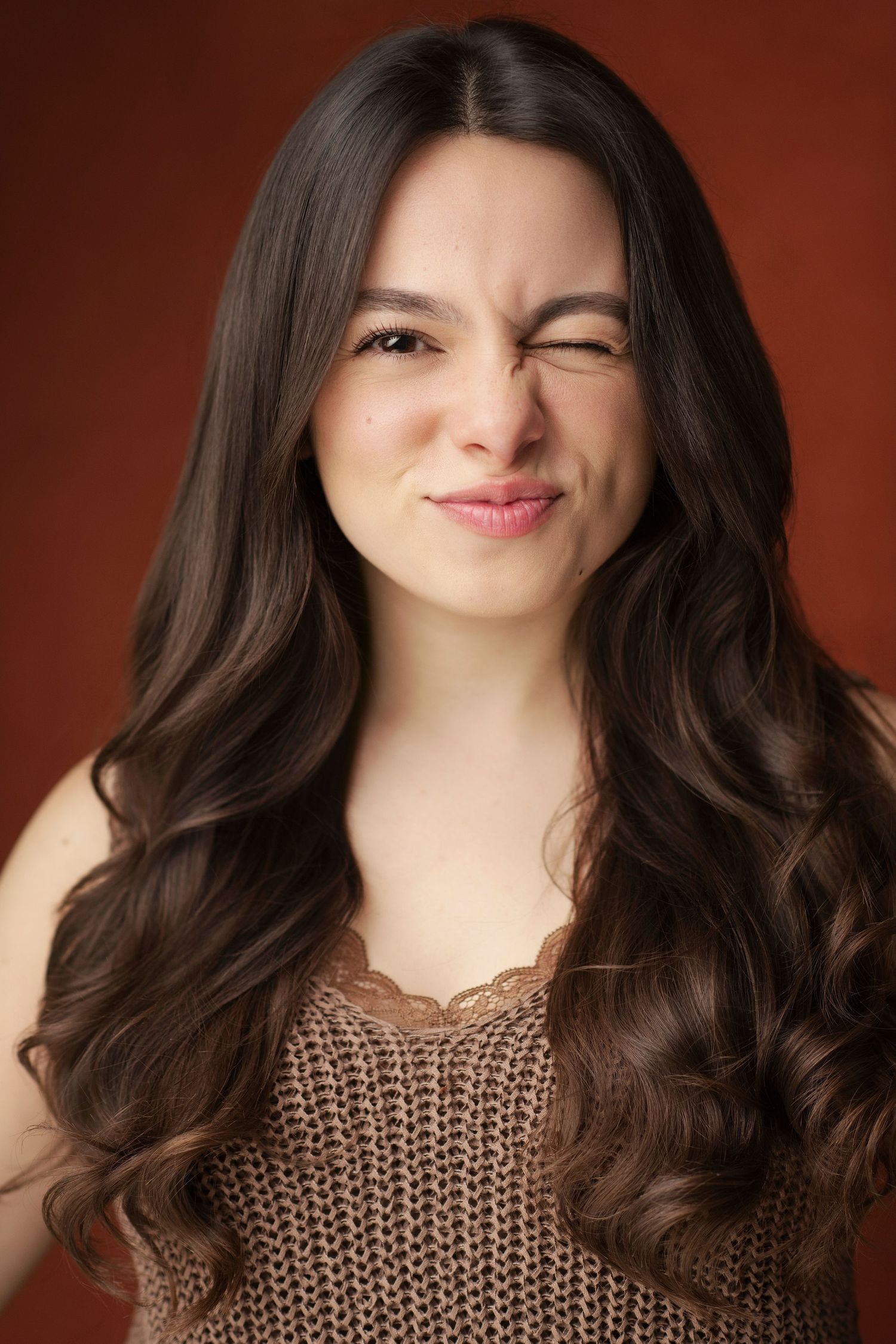 Young woman with long wavy brown hair winking playfully, wearing a brown knit top against a rust background.