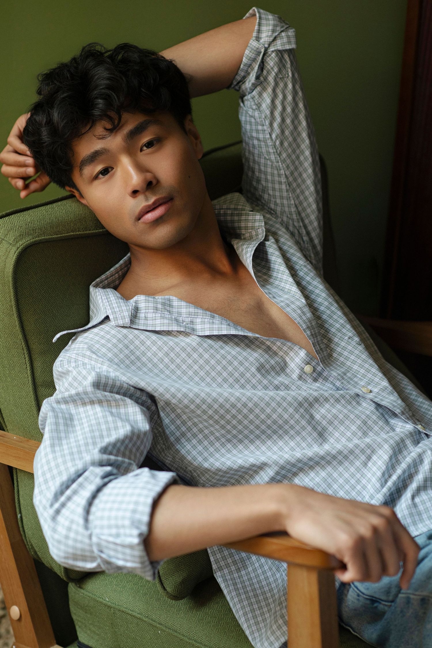 Young man with dark curly hair lounges in a green vintage armchair wearing an unbuttoned light blue checkered shirt and jeans.
