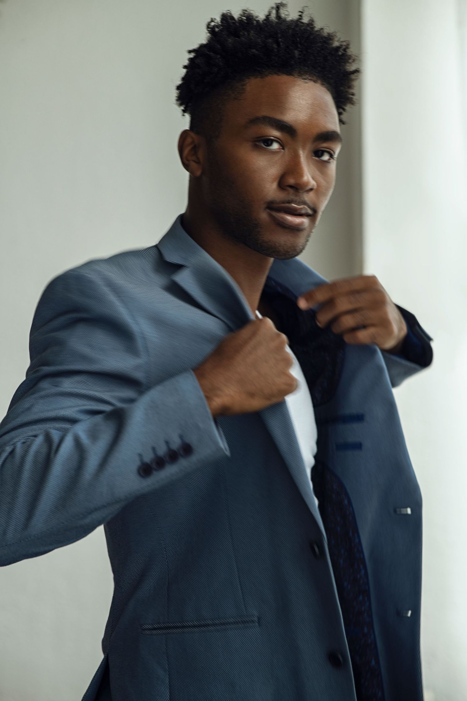Young Black man in a blue suit jacket adjusting his lapels, posing against a light gray background.