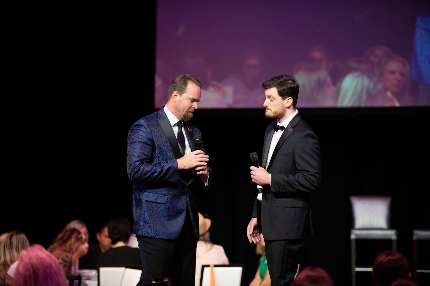 Two men in formal suits speaking on stage at an elegant gala event with audience seated in background.
