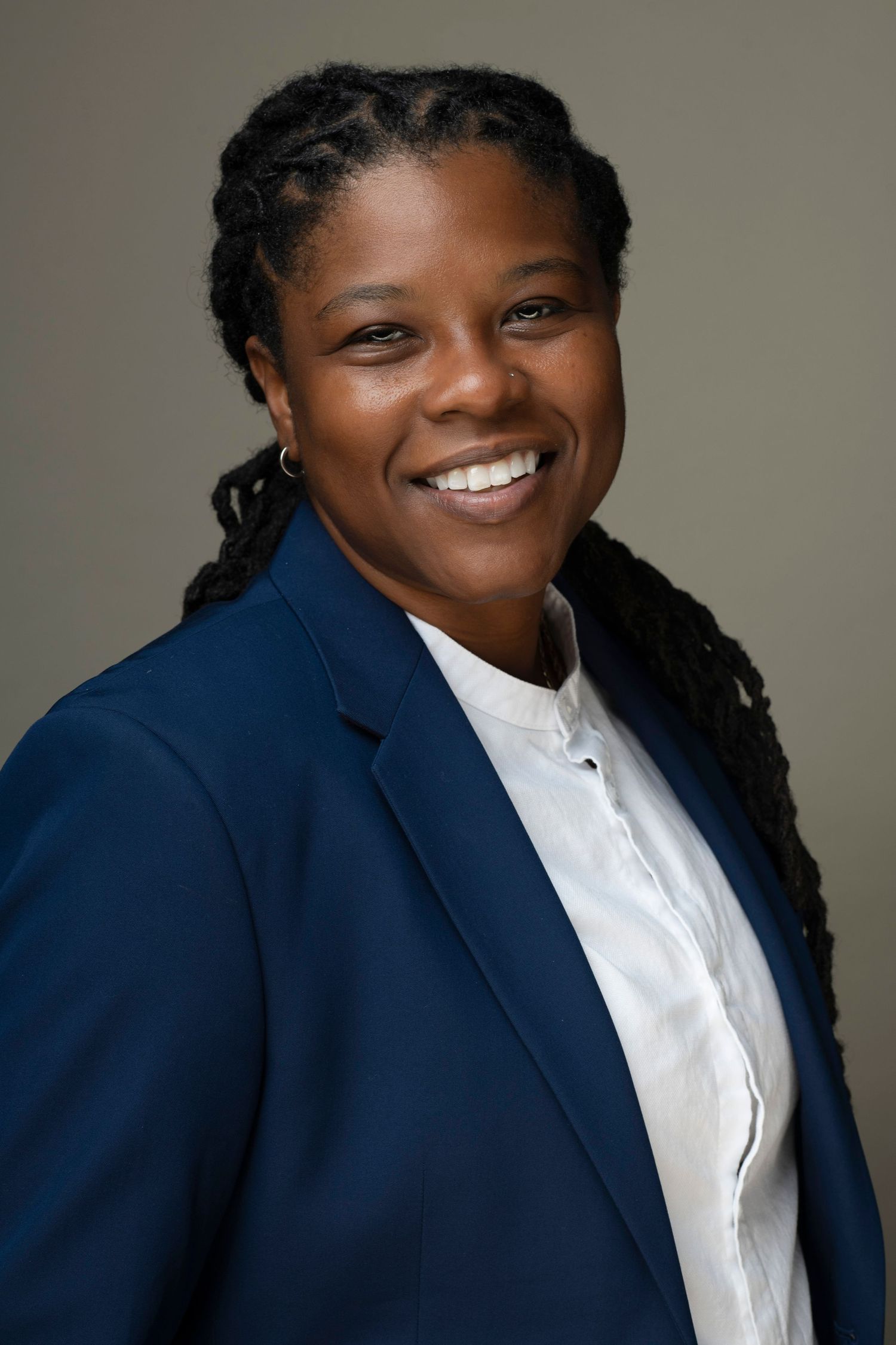 Professional woman in navy blazer and white shirt smiling confidently, with braided hair, against neutral background.