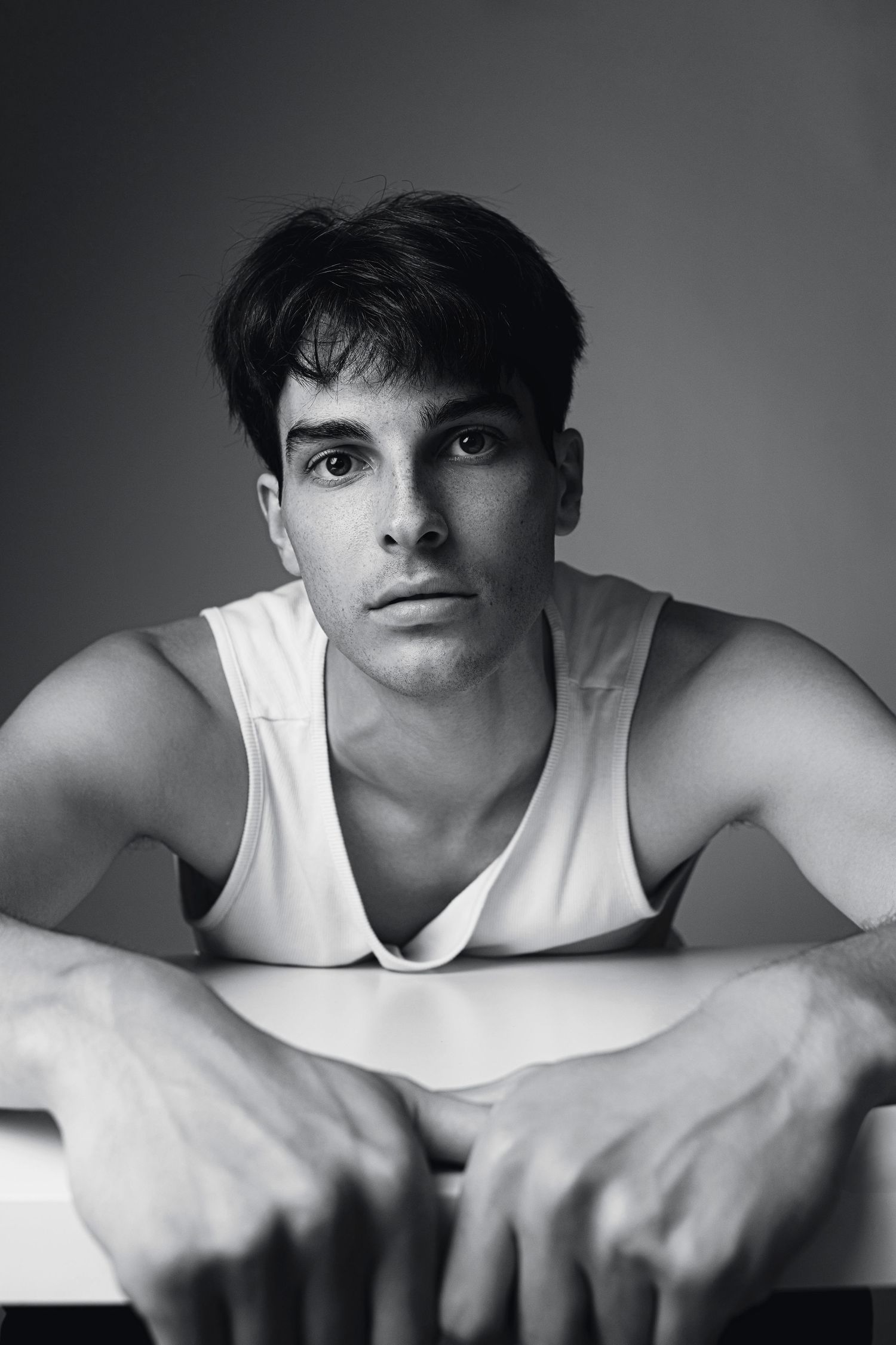 Black and white portrait of a young man in a white tank top leaning on a surface.