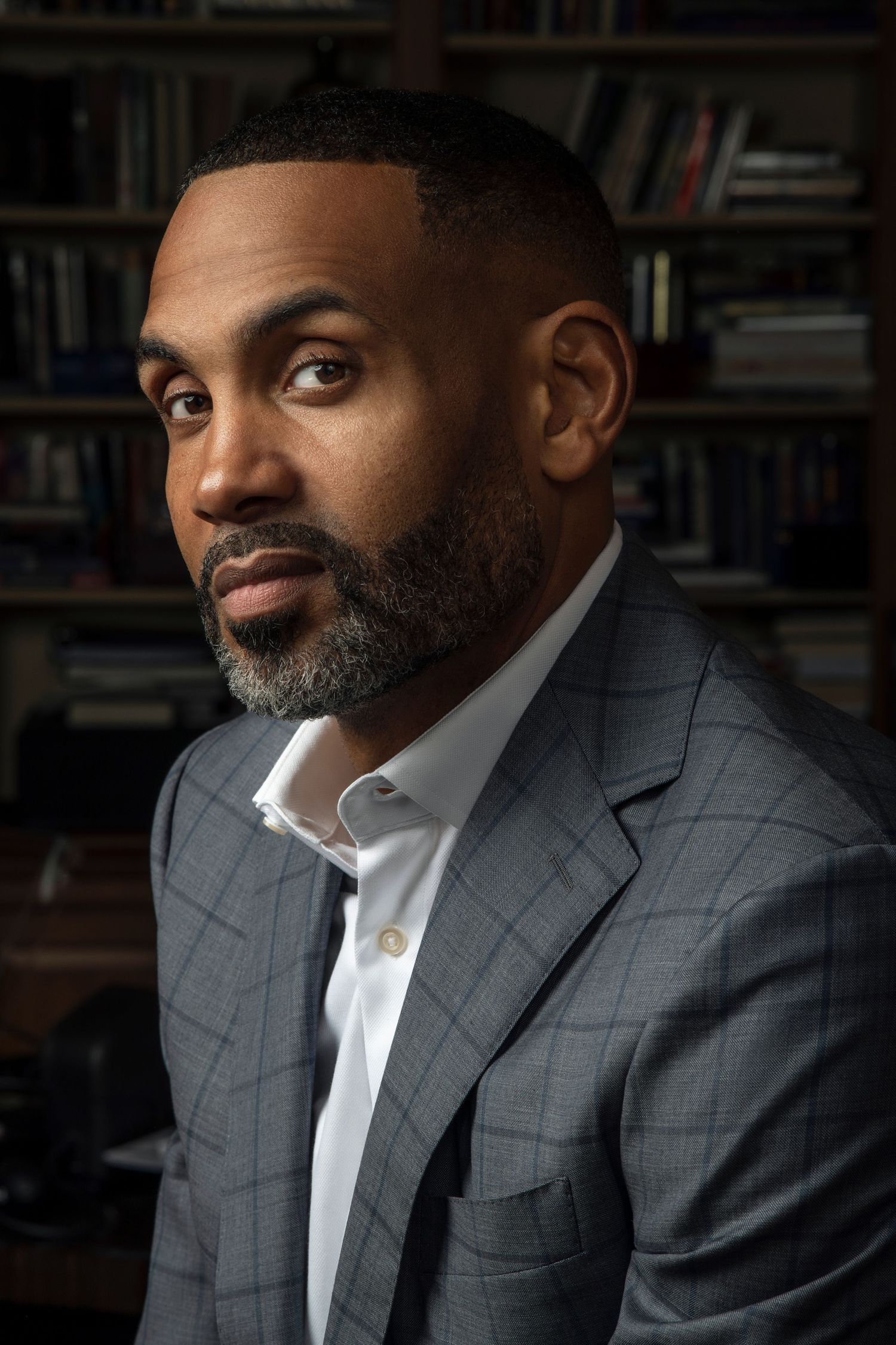 Man with a trimmed salt-and-pepper beard wearing a gray plaid blazer and open-collar white shirt, seated in front of filled bookshelves.