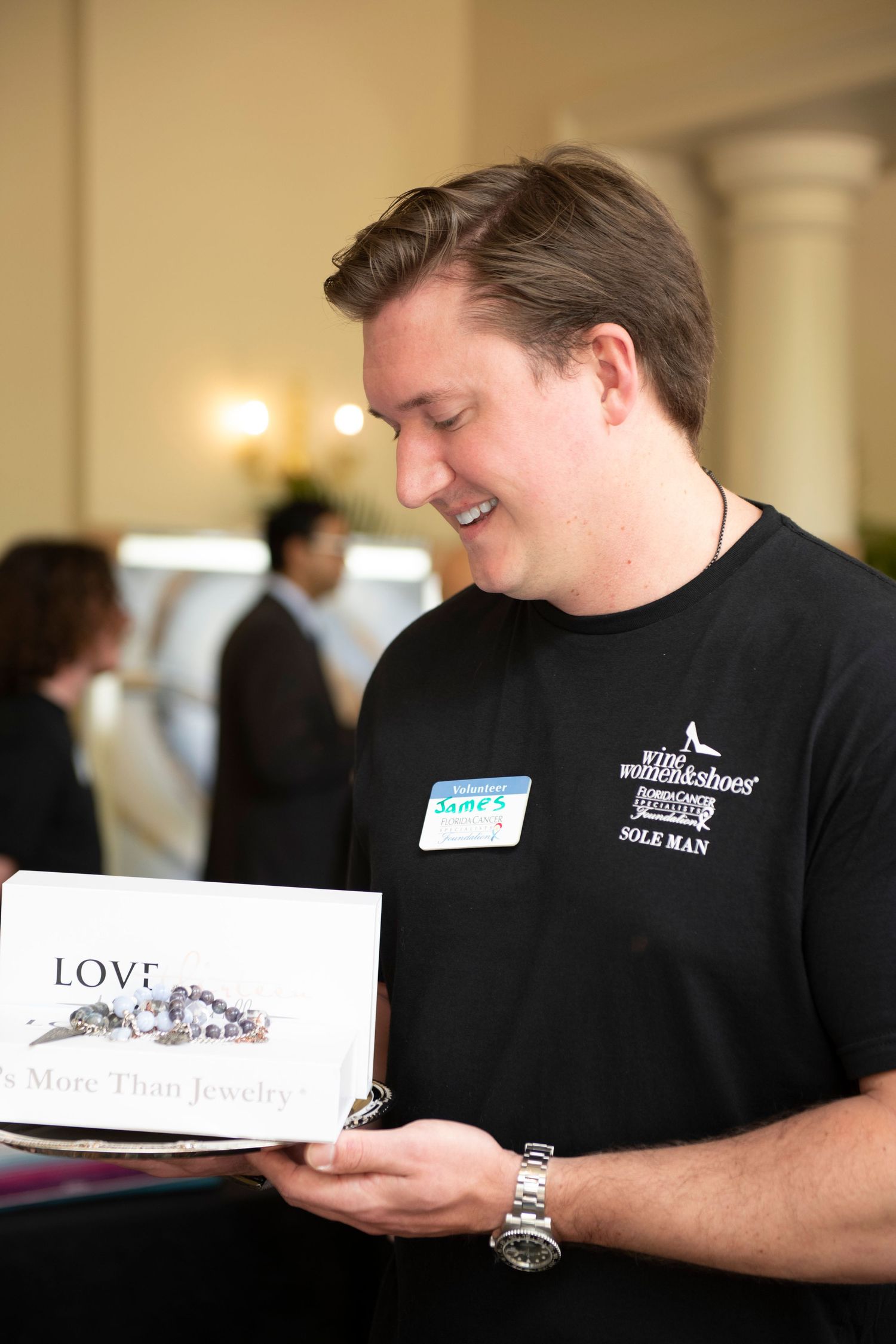 Man in black Wikishoes t-shirt smiling while reading a jewelry brochure at an indoor event.
