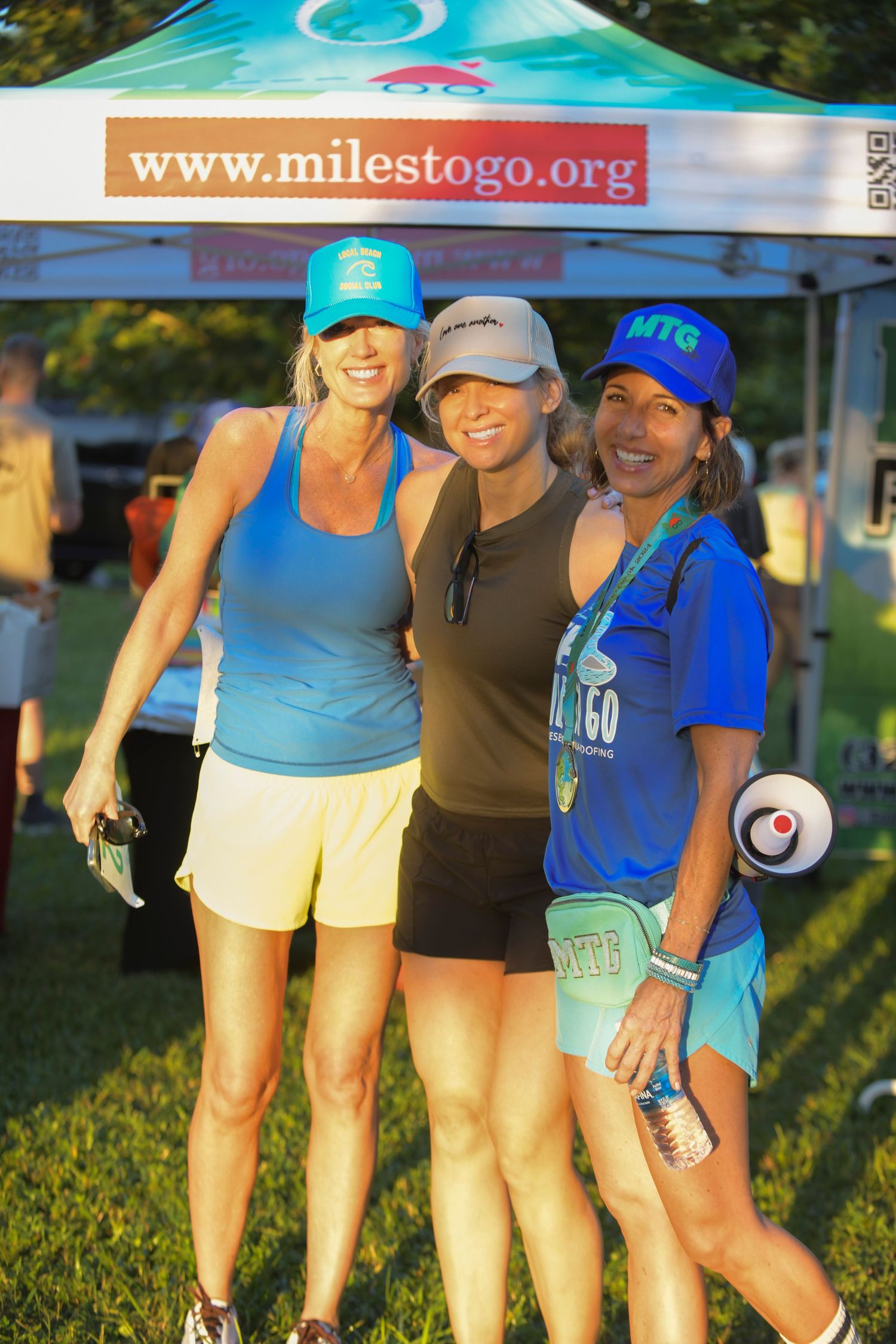 Three women pose together at a Miles to Go charity running event outdoors, wearing athletic gear and baseball caps.
