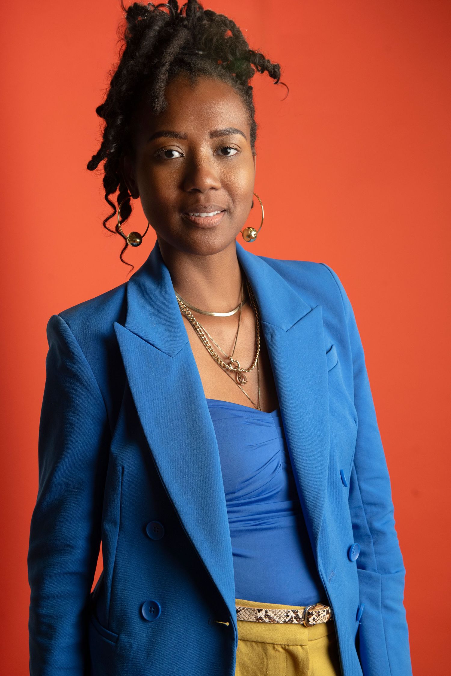 A young Black woman in a blue blazer and yellow skirt poses confidently against a vibrant red background.