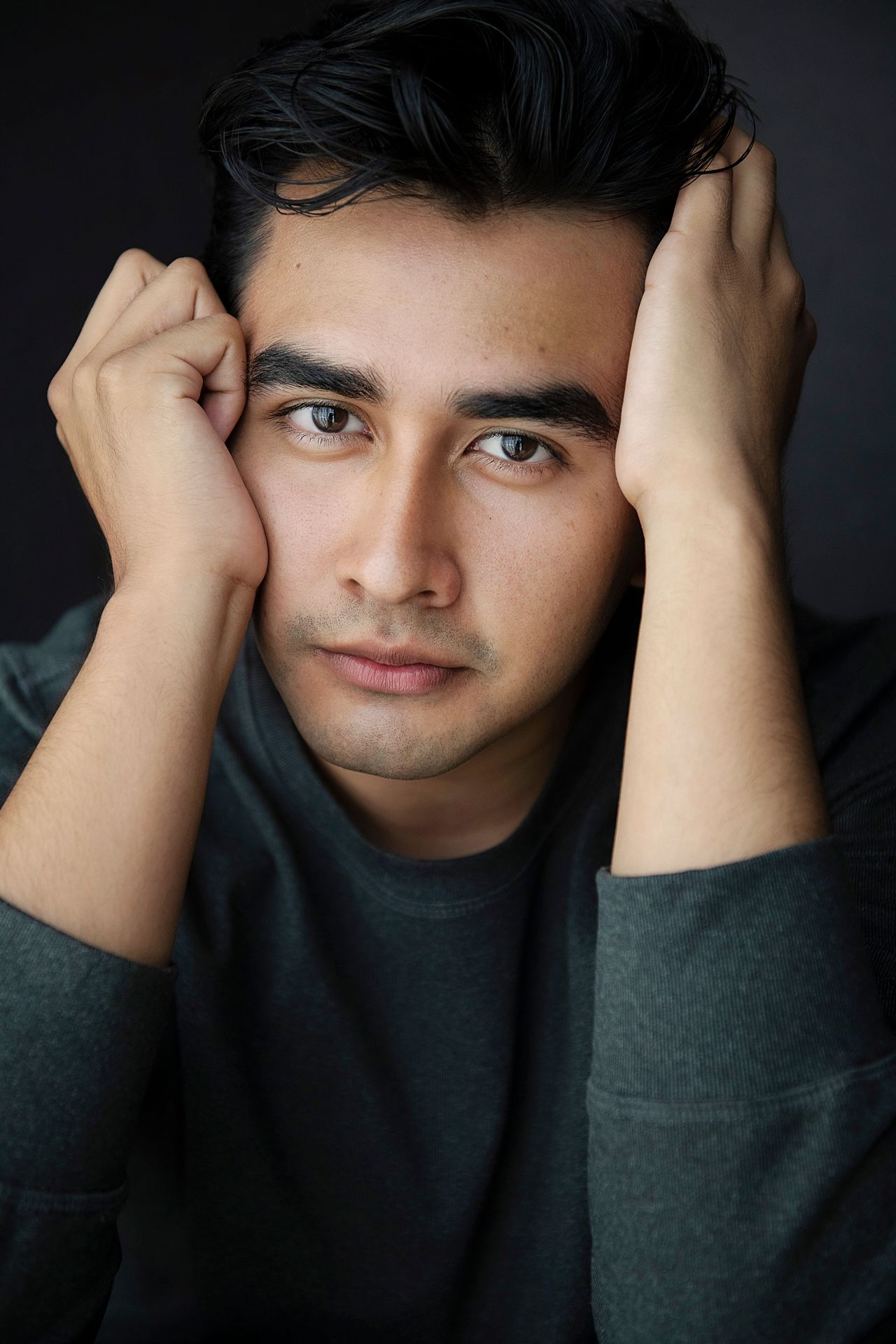 Young man with dark hair rests his hands on his head in a moody dark studio headshot.