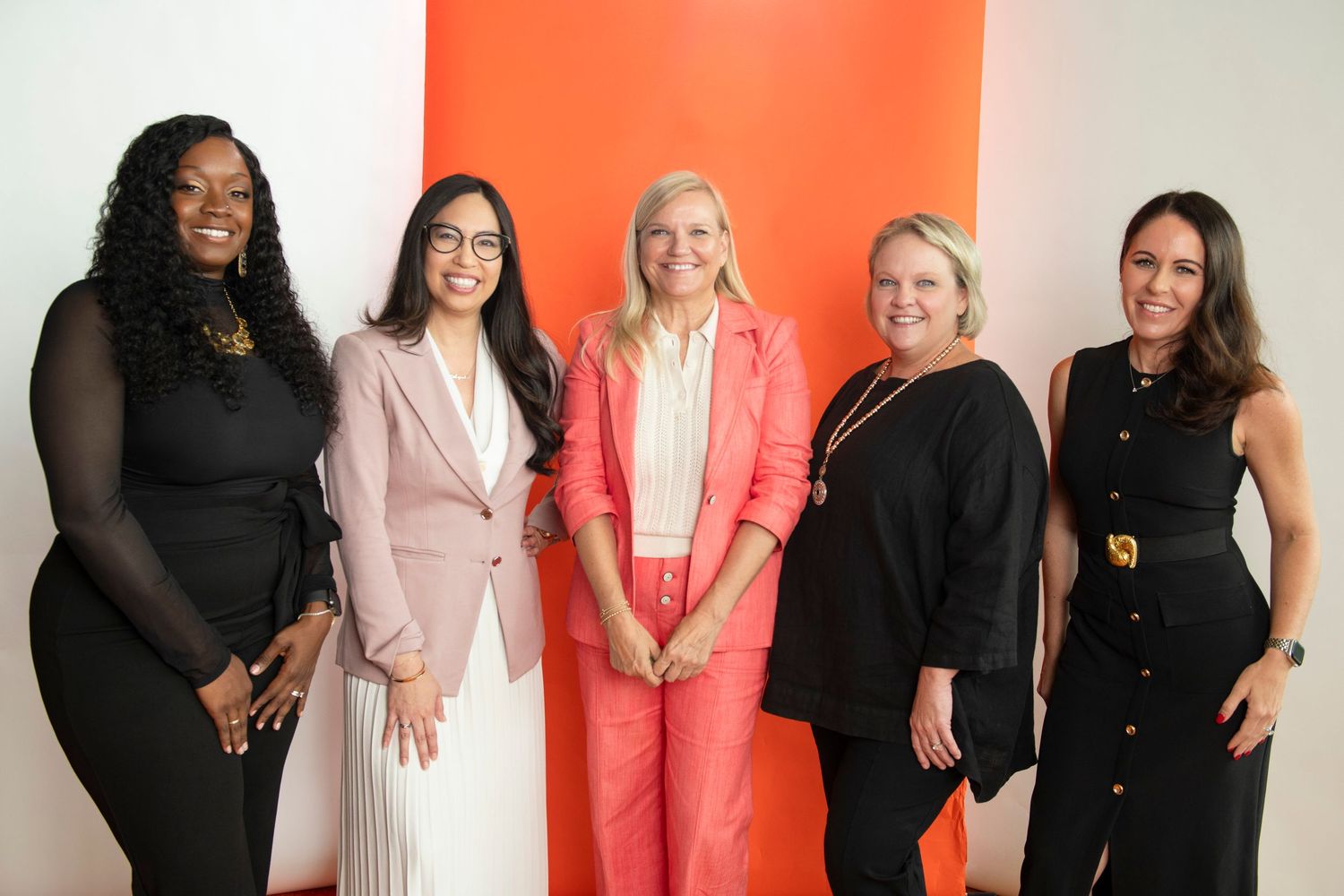 Five professional women smiling together in front of a bright orange wall in a modern office setting.
