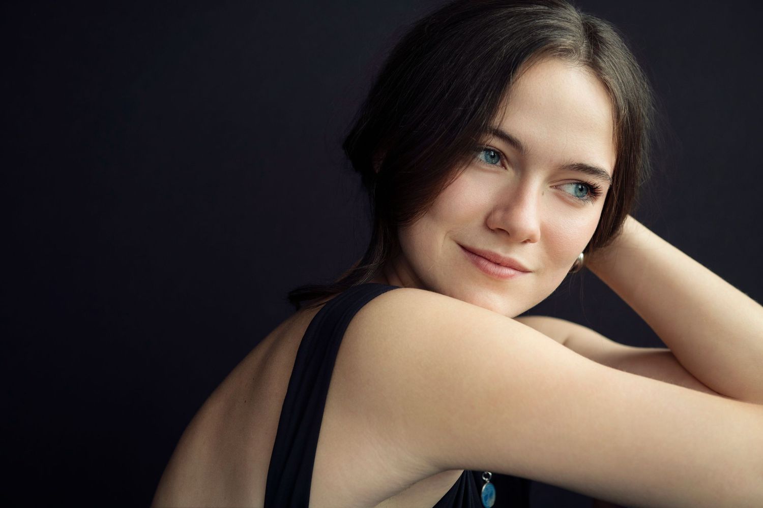 Young brunette woman with blue eyes posing with arms crossed, smiling against dark background.