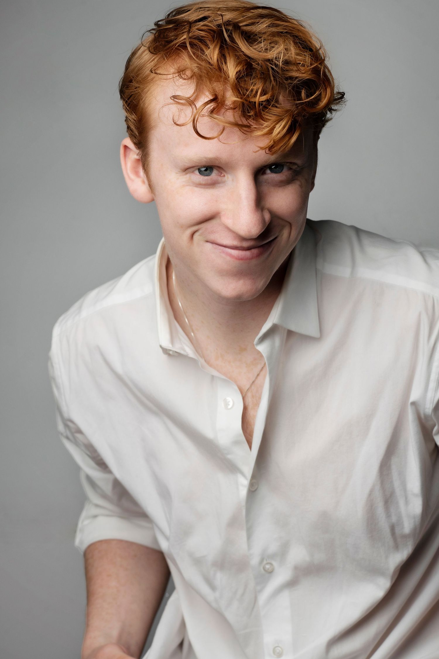 Young man with red curly hair smiles wearing white button-up shirt against gray background.