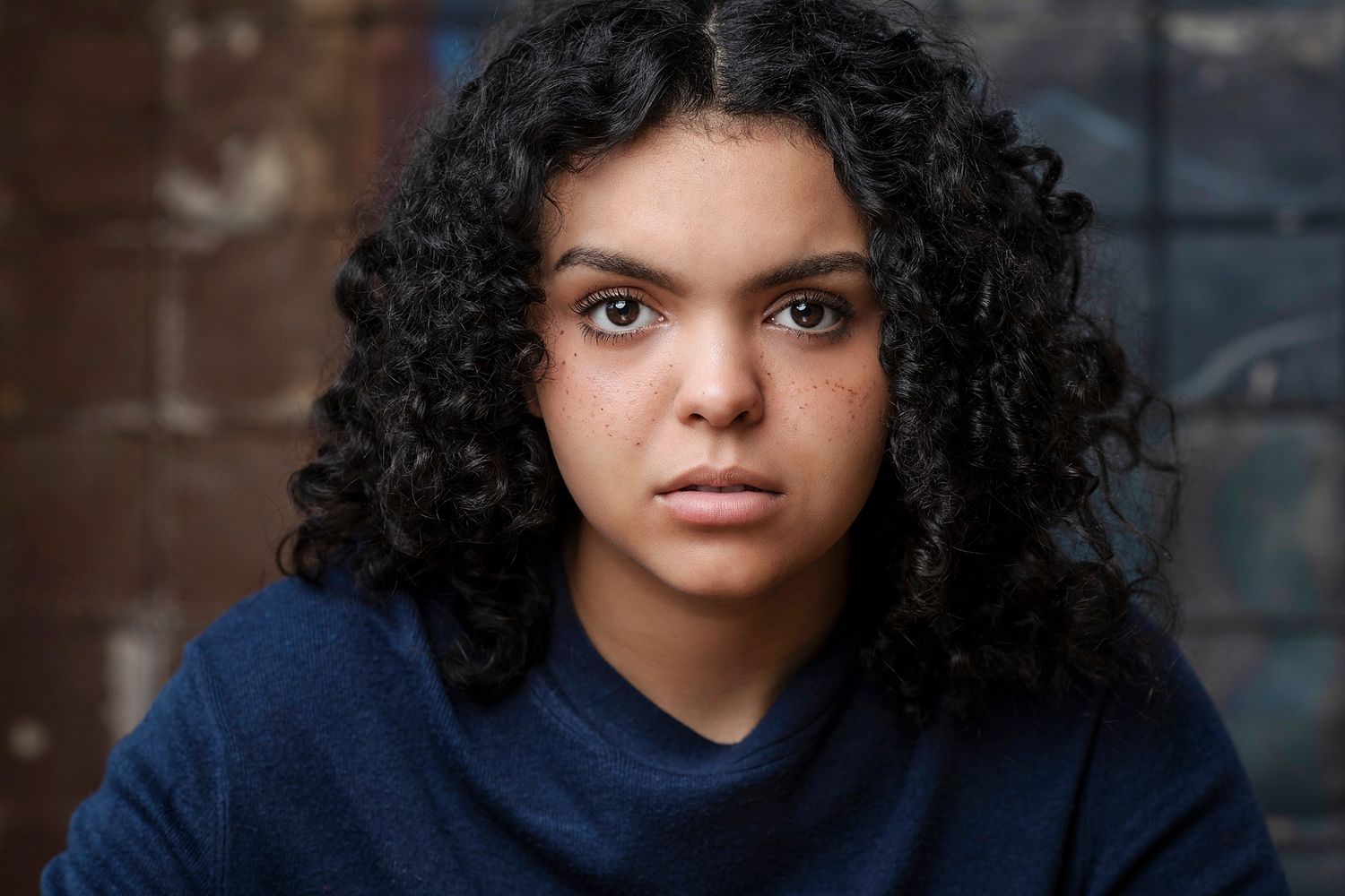 Young mixed-race woman with curly black hair wearing a navy sweater against a brick background.
