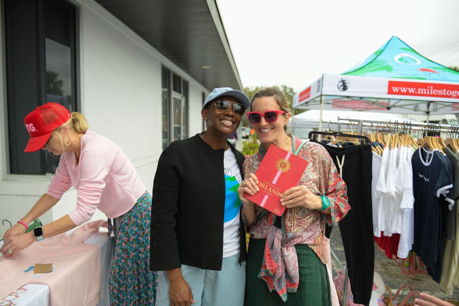 Two people smiling at an outdoor market event, with clothing racks and a green vendor tent visible in the background.