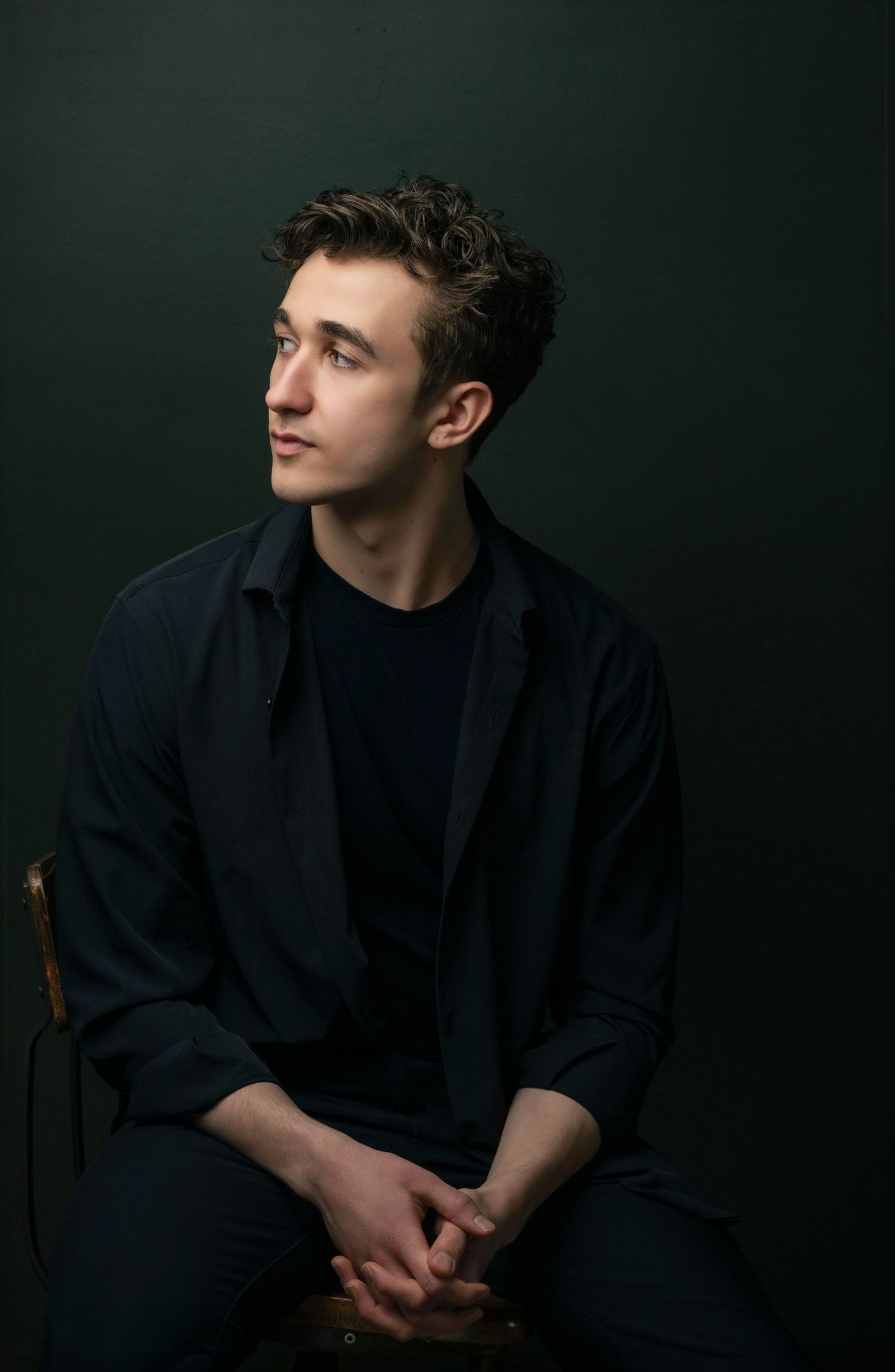 Young man with tousled dark hair in a black jacket and shirt sits with clasped hands, gazing off to the side against a dark backdrop.