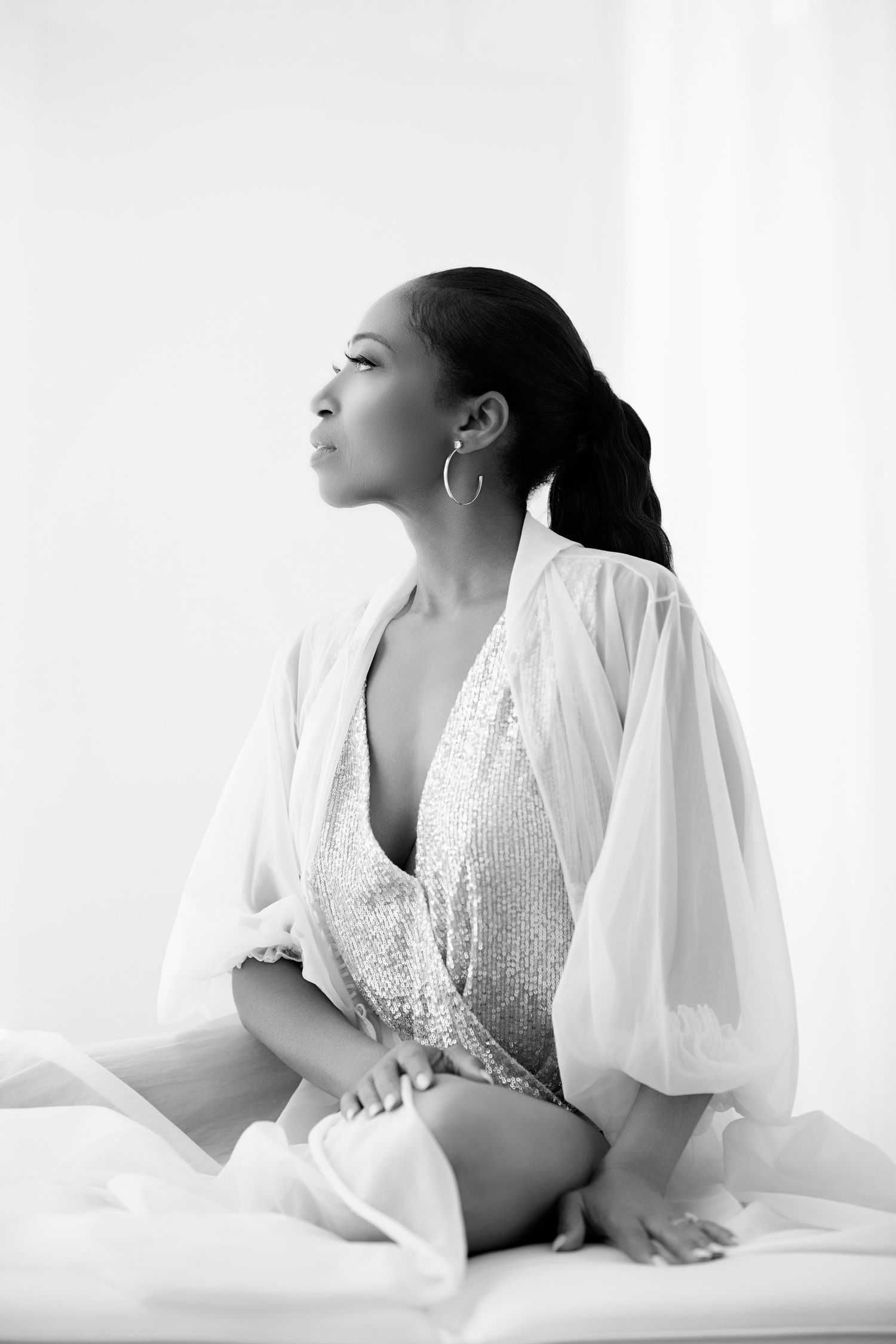 Black and white photo of a woman in sequined bodysuit and sheer robe sitting gracefully in soft light.