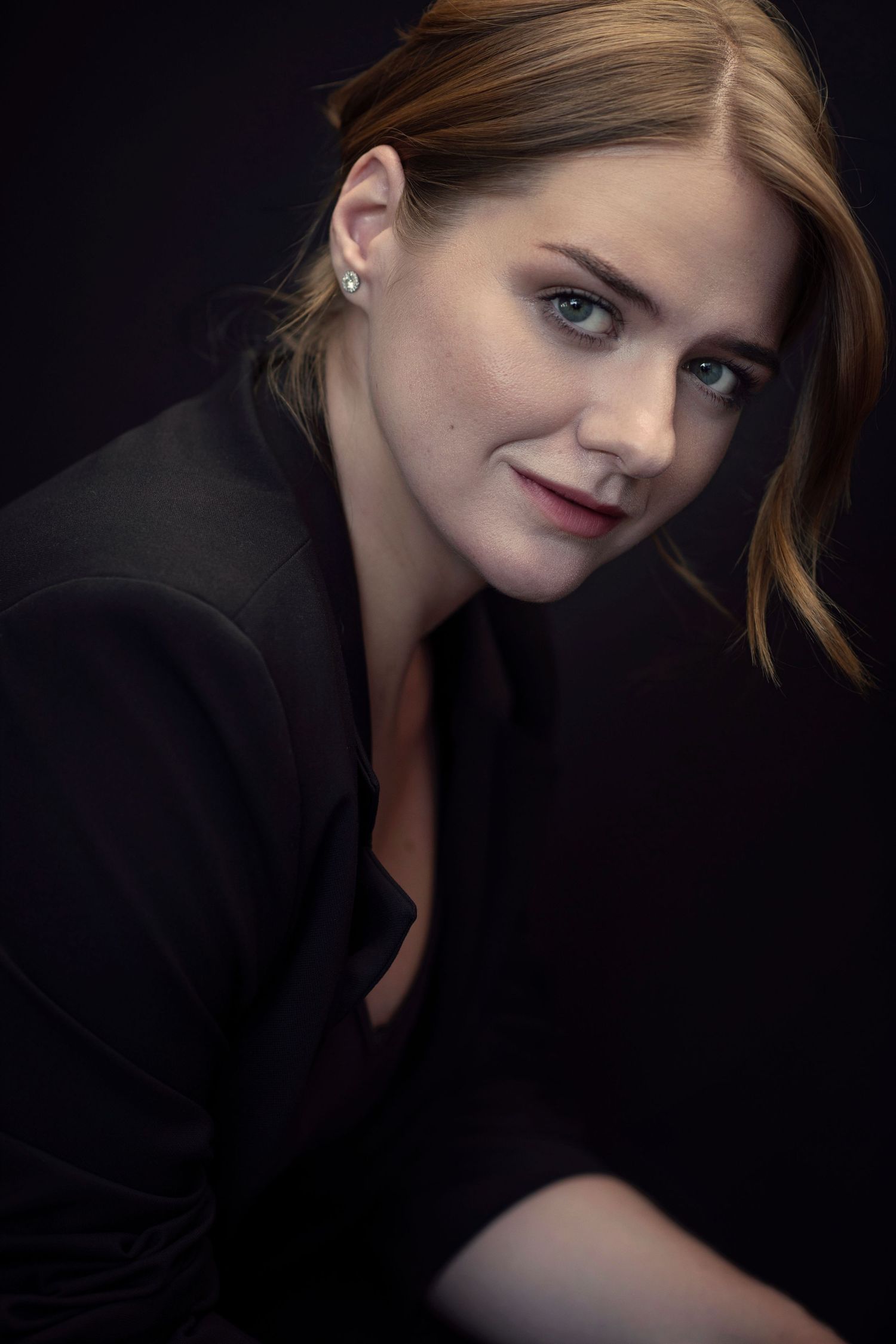 Portrait of a woman with auburn hair, blue eyes and stud earrings, wearing a black blazer and smiling softly against a dark background.