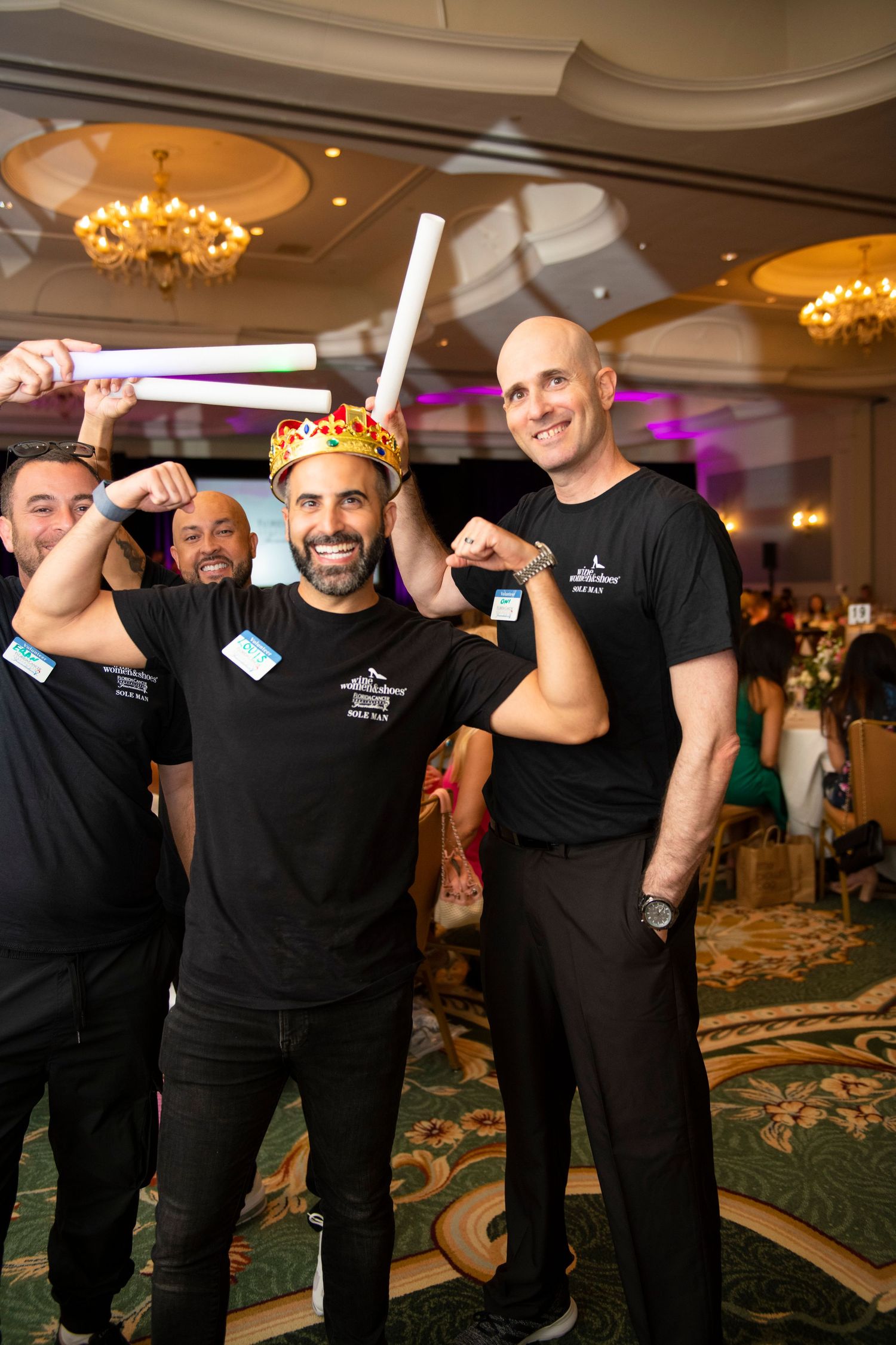 Men in black t-shirts celebrate at a gala event, one wearing a crown and holding glow sticks in a ballroom.