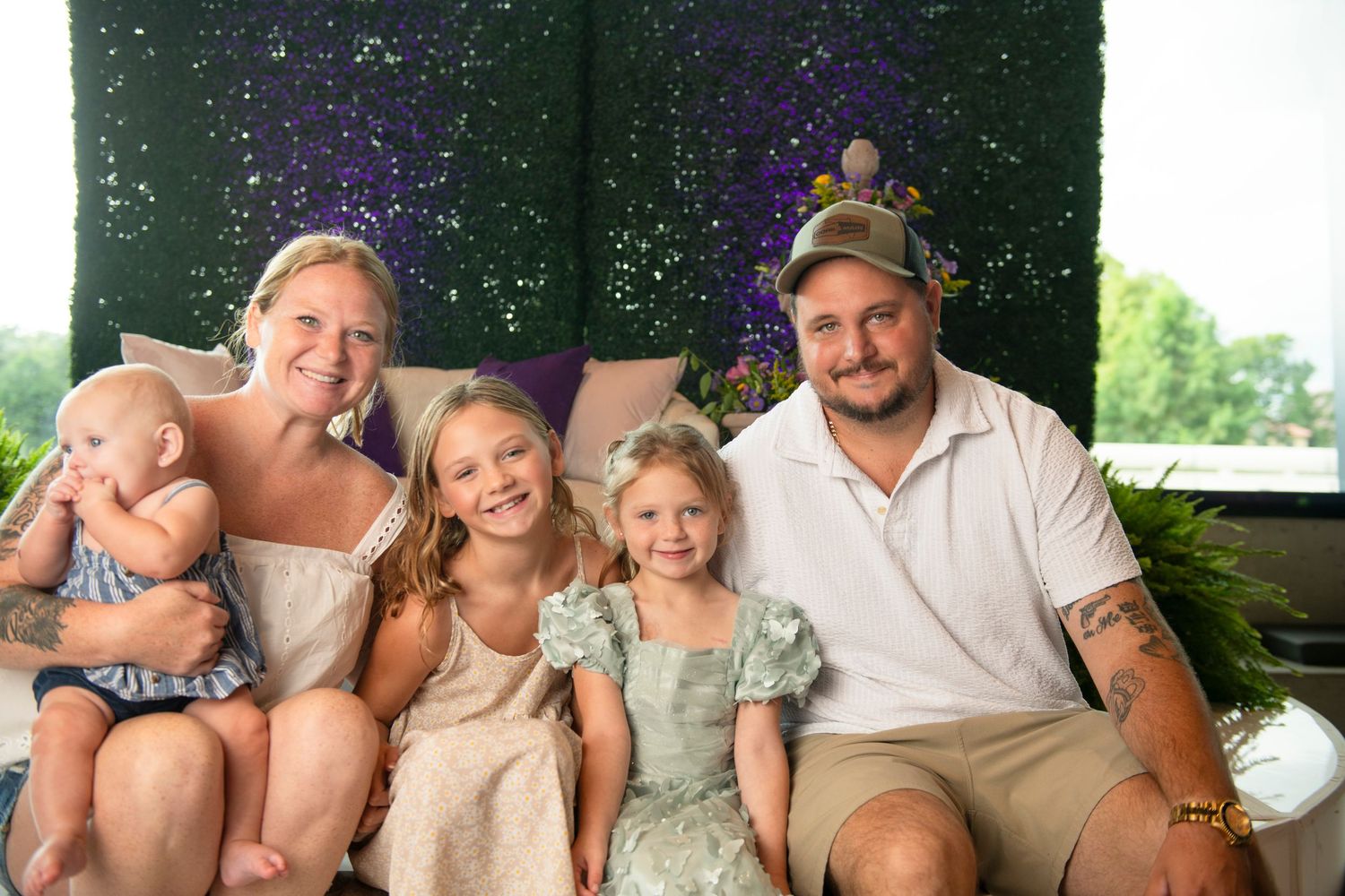 Happy family of five posing together outdoors, with parents and three young daughters smiling in front of a floral backdrop.
