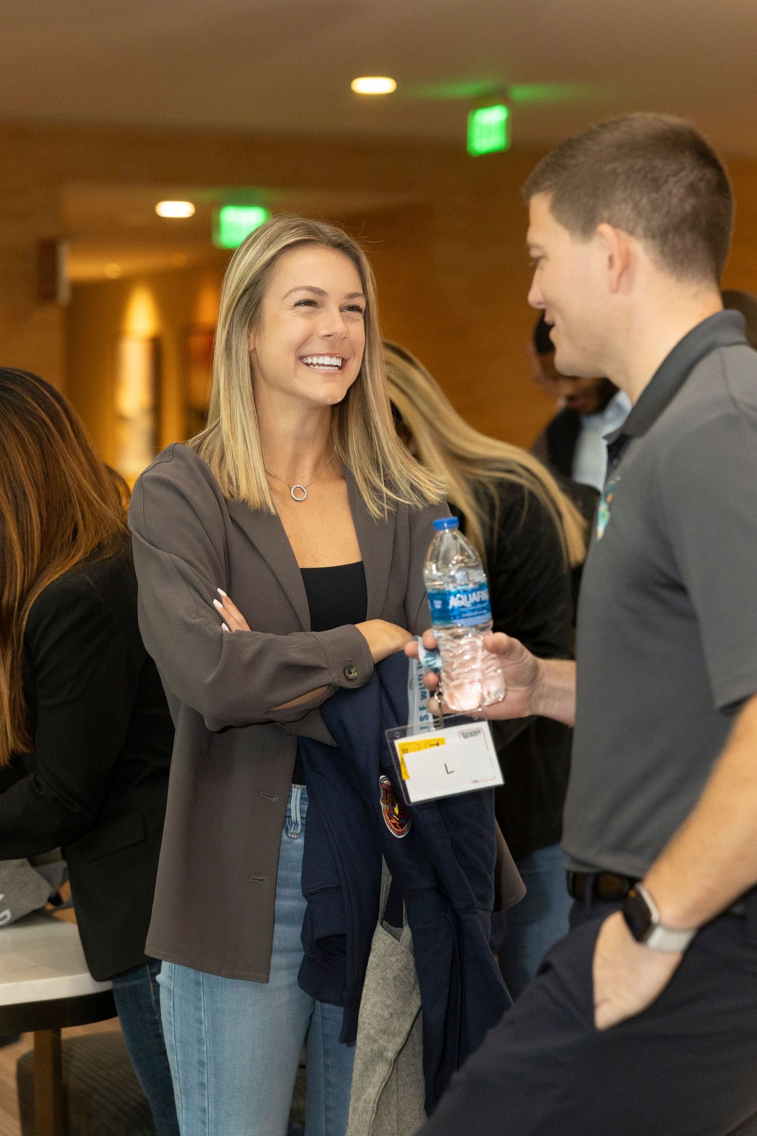 A smiling blonde woman in a blazer chats with a man holding a water bottle at a professional networking event.