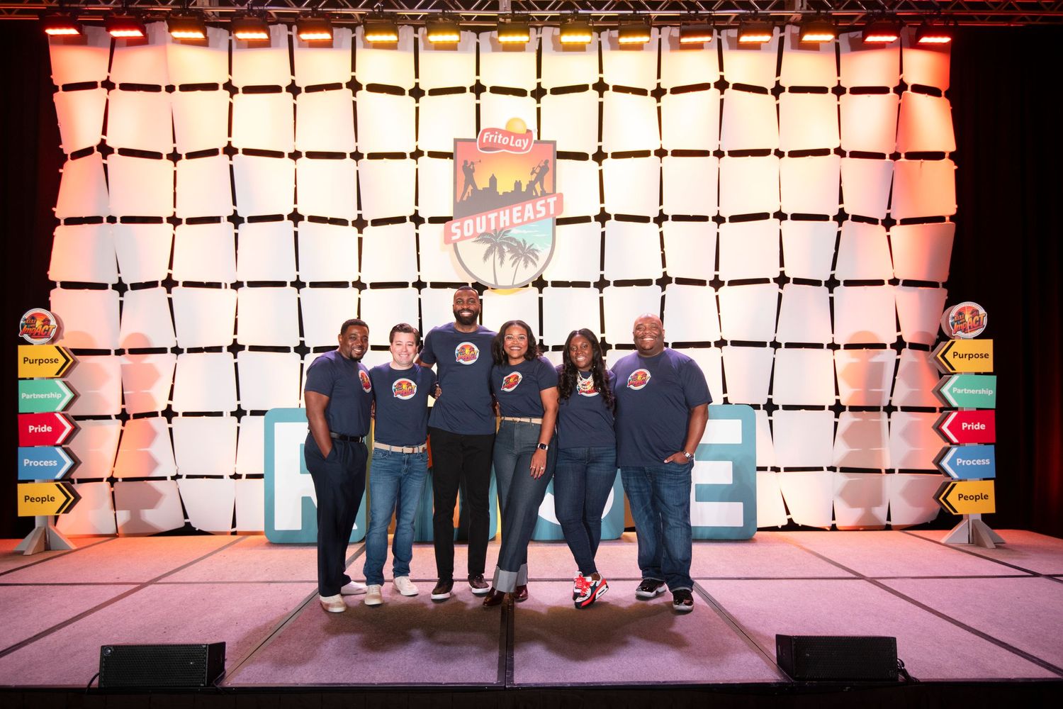 Group of five people in matching blue shirts posing on stage in front of a colorful illuminated backdrop.