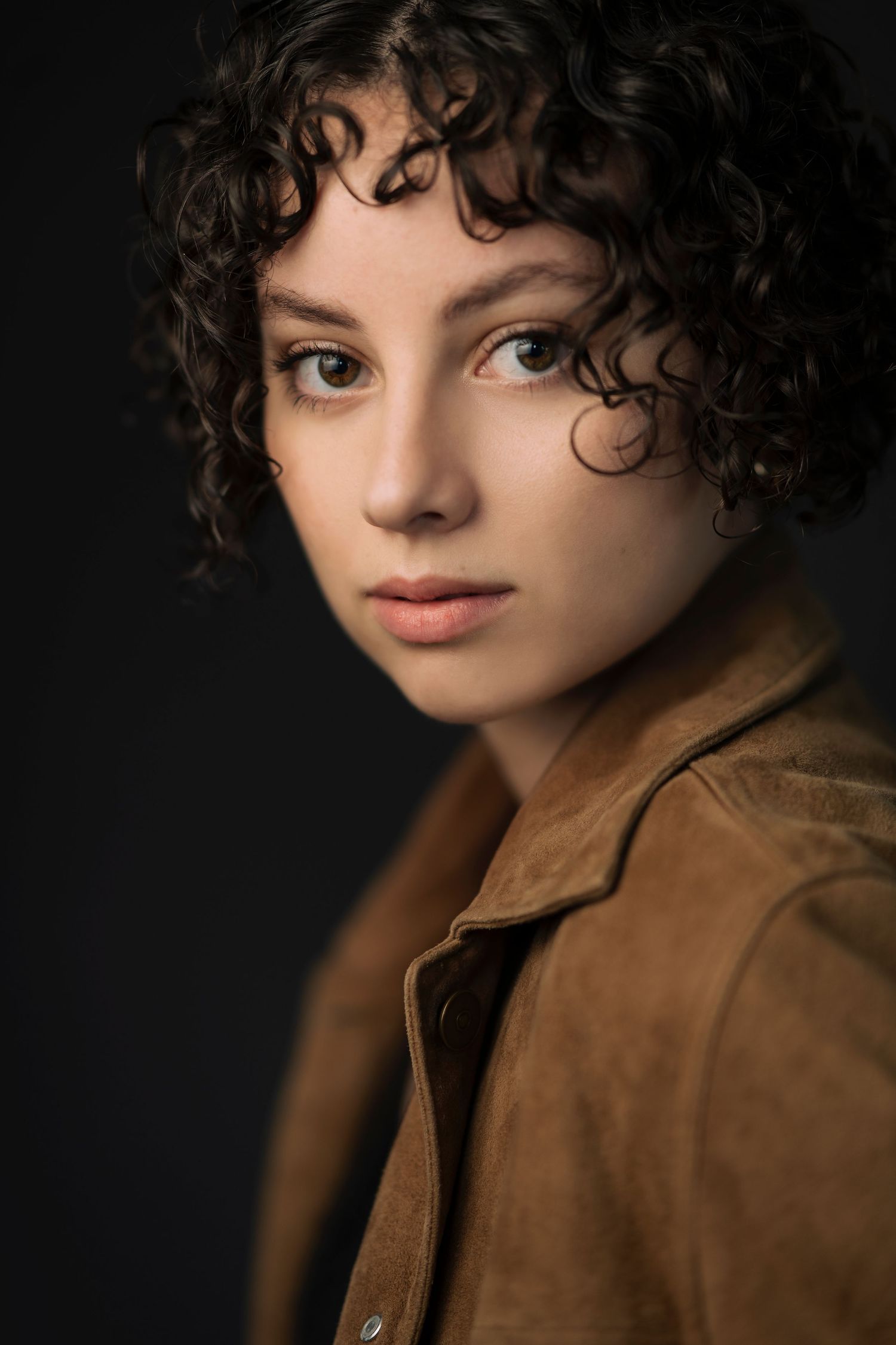 Portrait of a young woman with short curly dark hair wearing a brown suede jacket against a dark background.