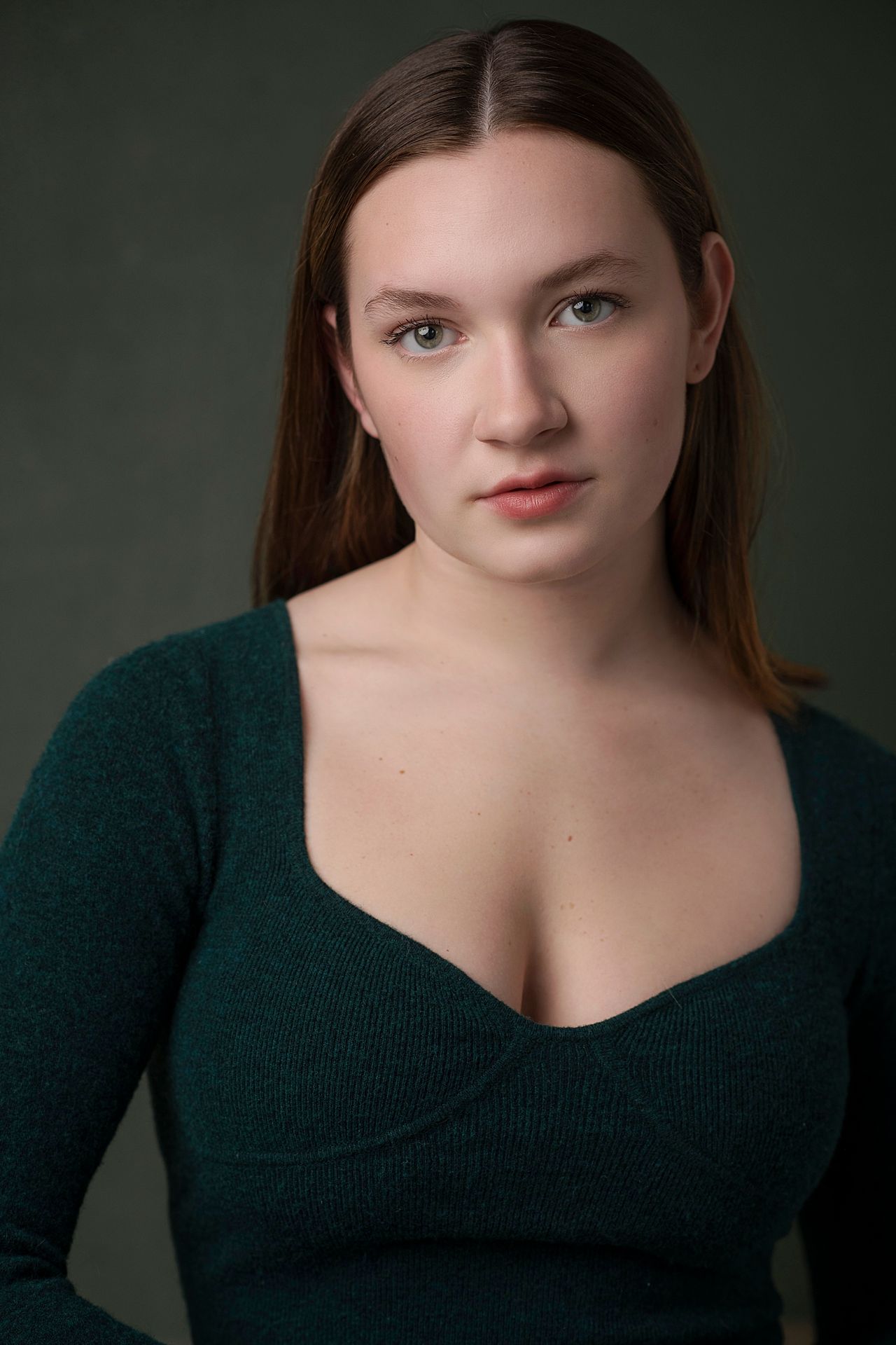 Young woman with straight brown hair wearing a teal V-neck sweater against a dark background.