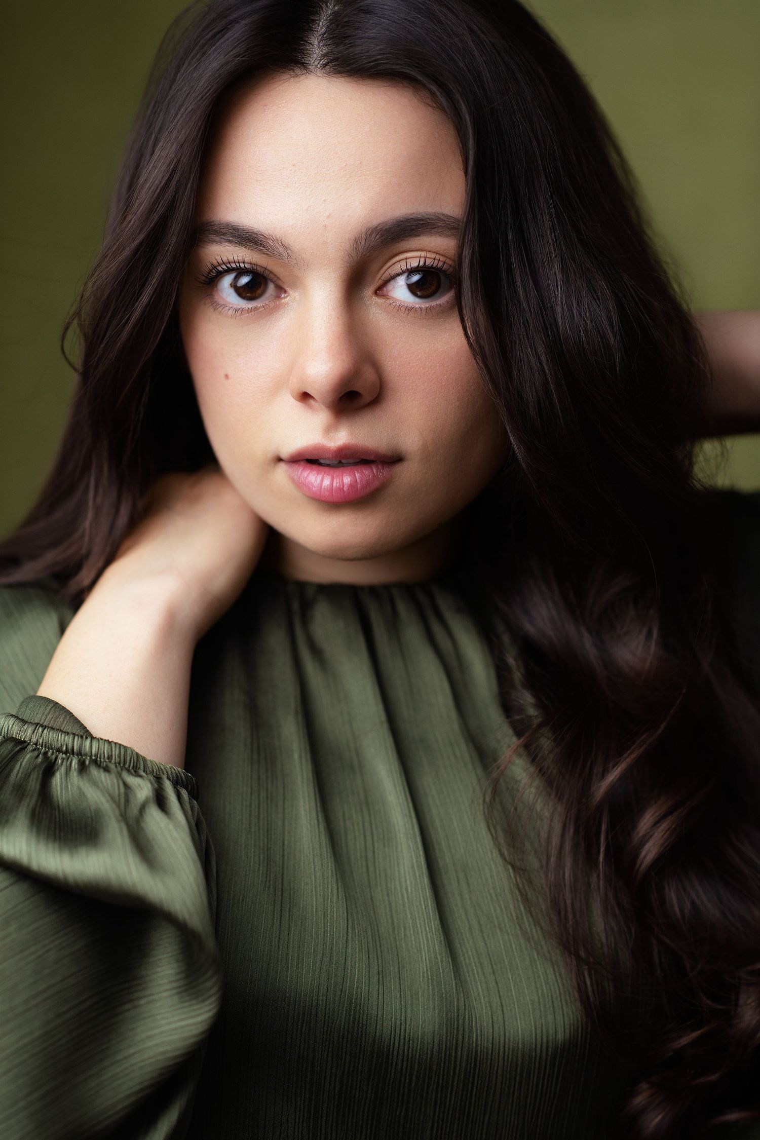 Portrait of a young woman with long wavy dark hair in an olive green satin blouse, hand resting behind her neck against a green backdrop.