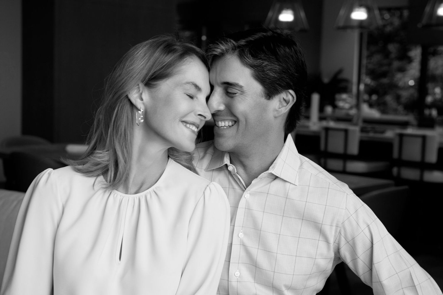 Black and white photo of a smiling couple leaning their heads together in a warm, affectionate moment inside a softly lit restaurant.