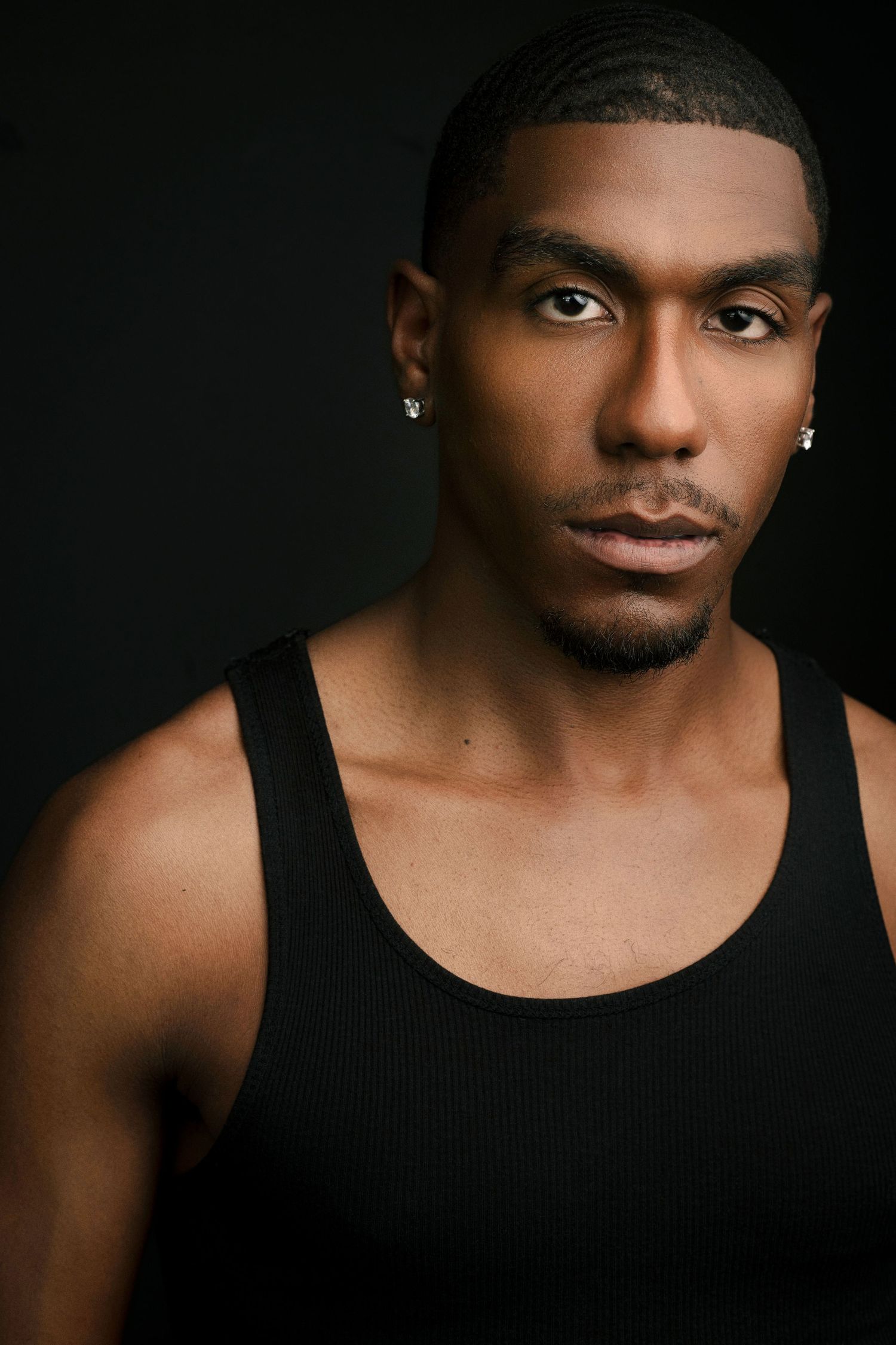 Studio portrait of a man with short hair, light stubble and stud earrings, wearing a black ribbed tank top against a dark background.
