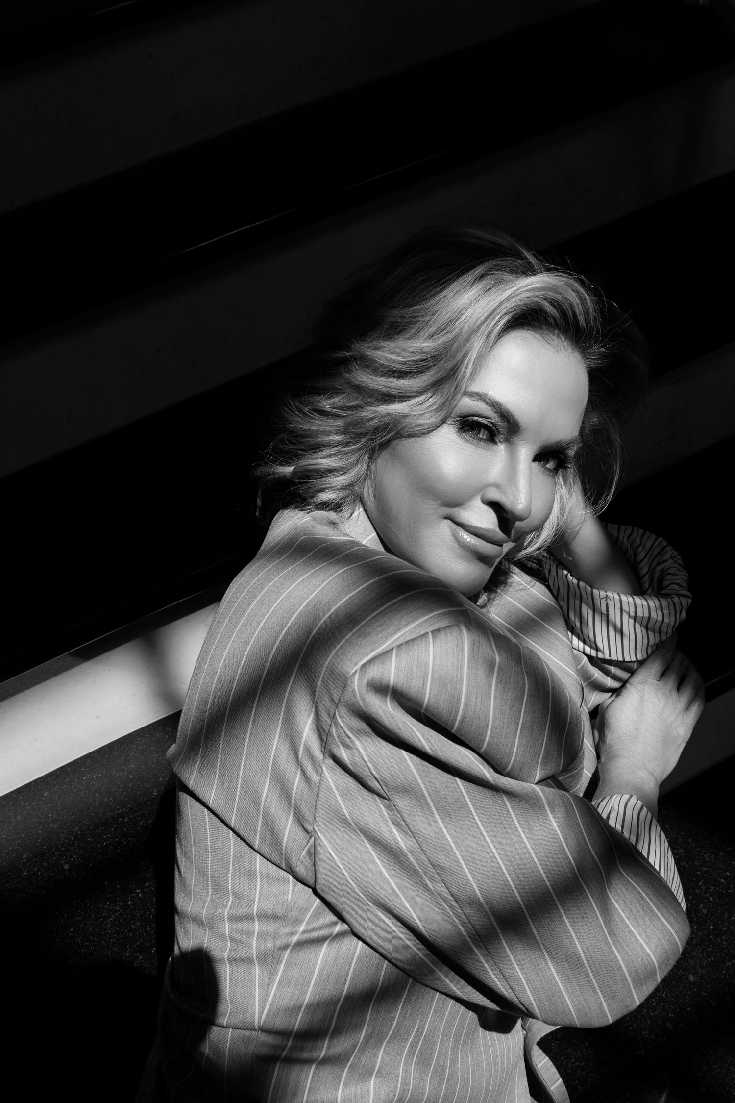 Black and white portrait of a smiling blonde woman in a striped blazer, posing elegantly with hand near face.
