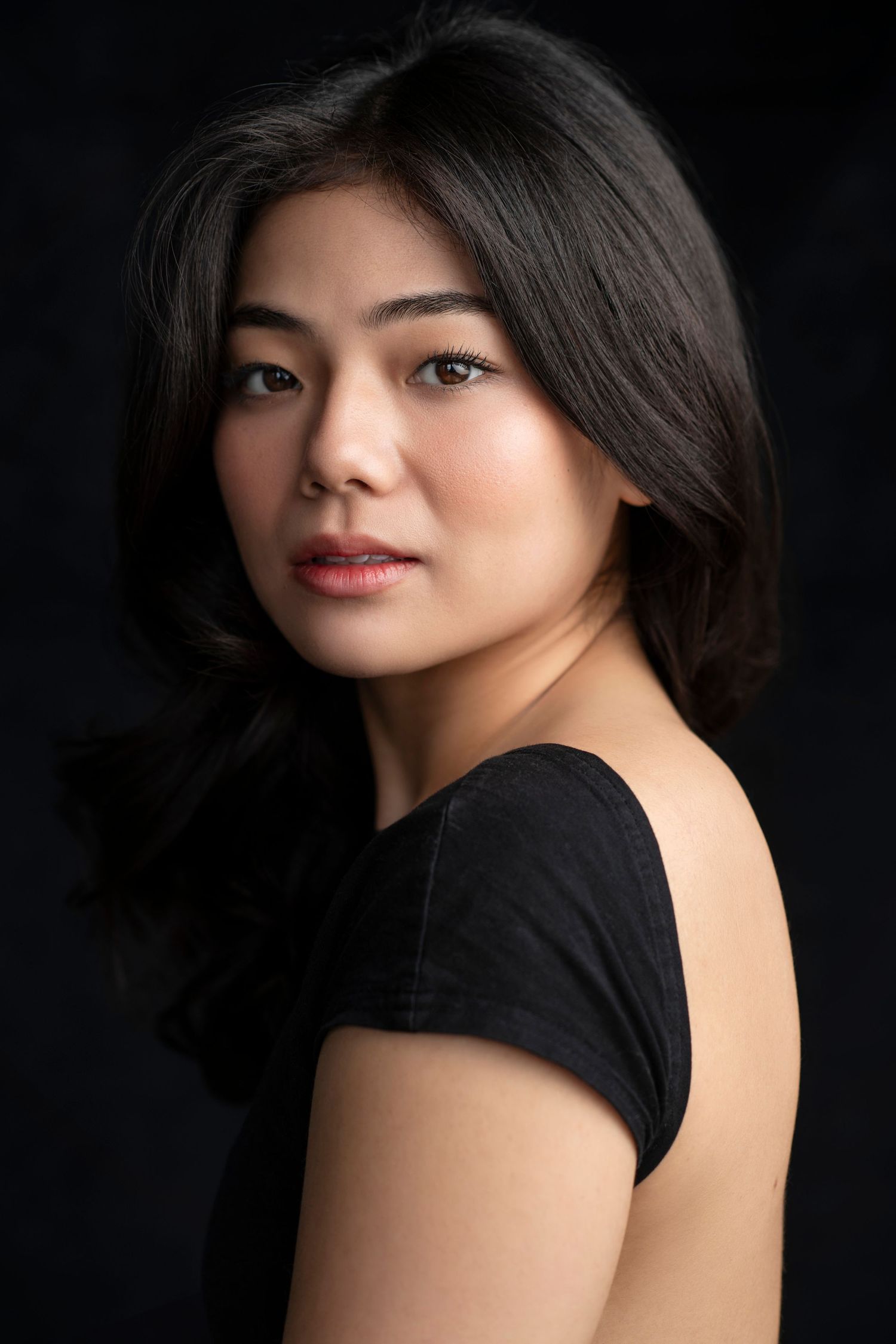 Asian woman with dark wavy hair wearing a black top against a dark background.