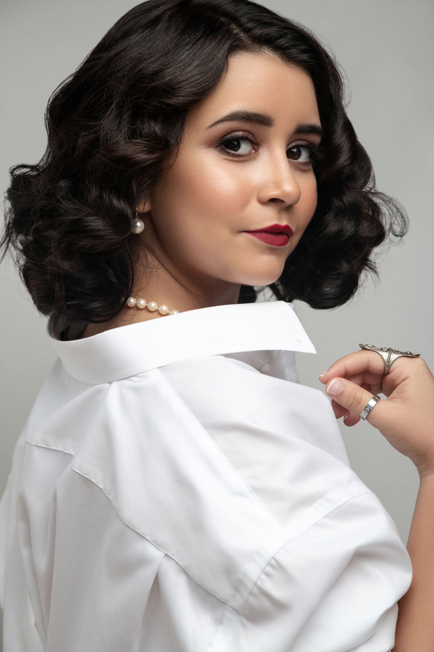 A young woman with dark curly hair wearing a white shirt and pearl necklace poses elegantly against a gray background.