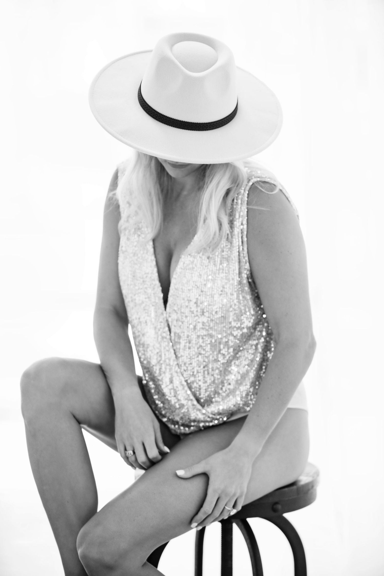 Black and white photo of a woman in a sequin top and wide-brim hat sitting on a stool.