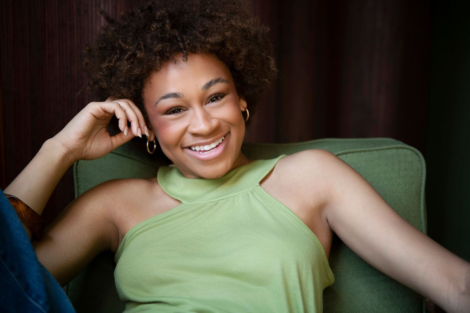 Smiling young woman with curly hair wearing a green halter top, relaxing in a green chair.