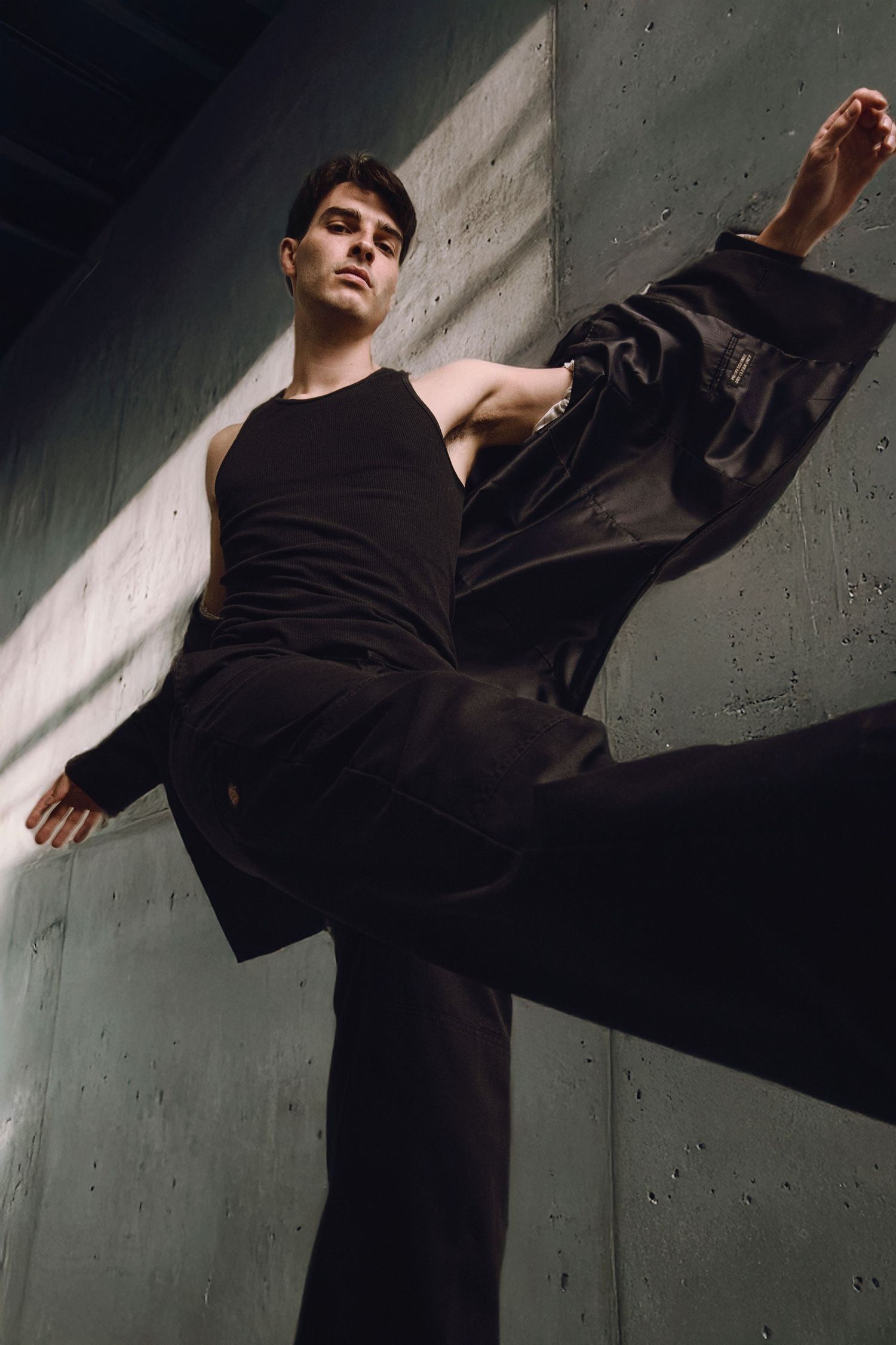 Male model in black outfit performs dynamic high kick pose against concrete wall.