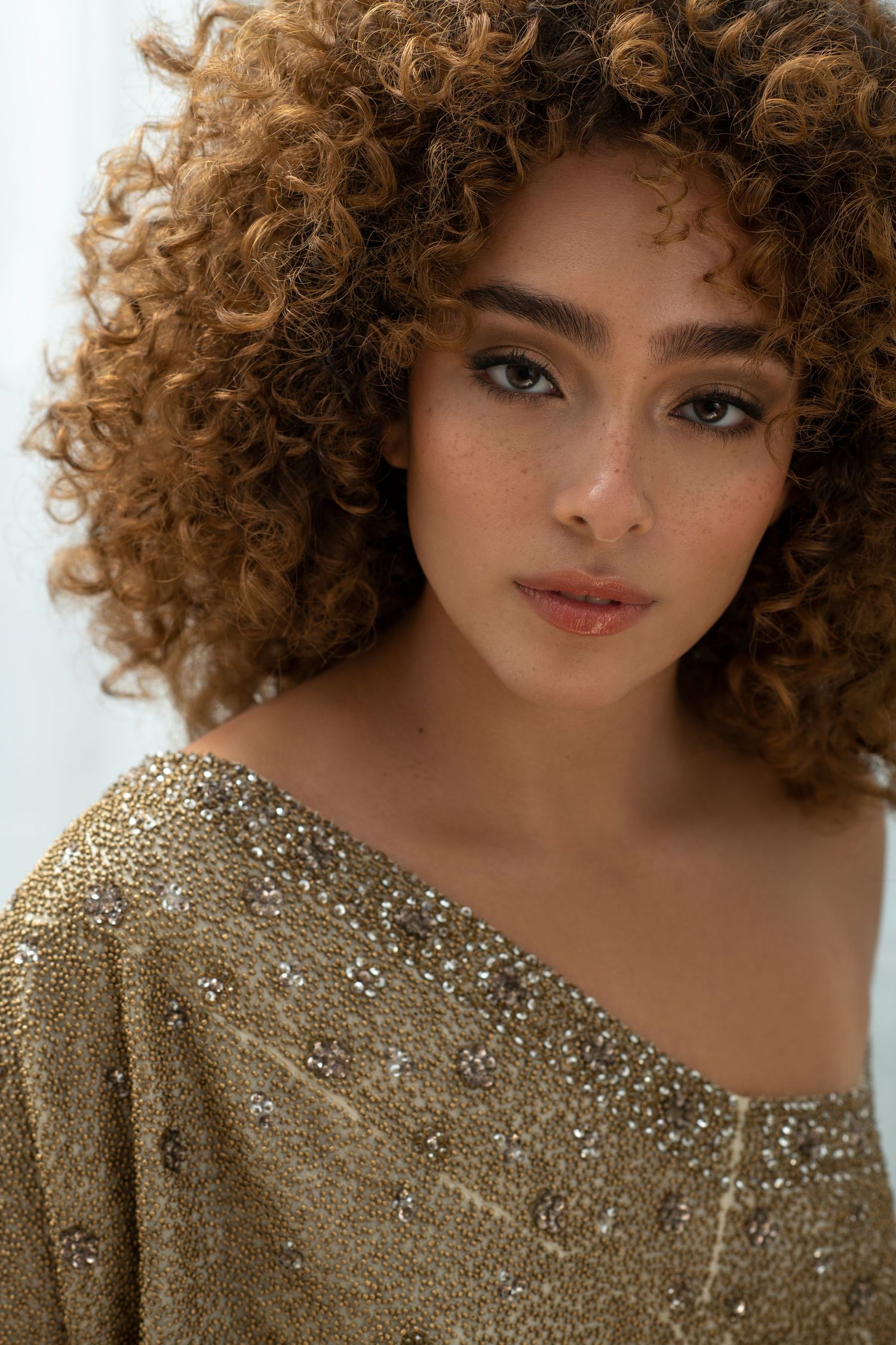 Young woman with curly auburn hair wearing a gold sequined off-shoulder top, posing elegantly for a fashion portrait.