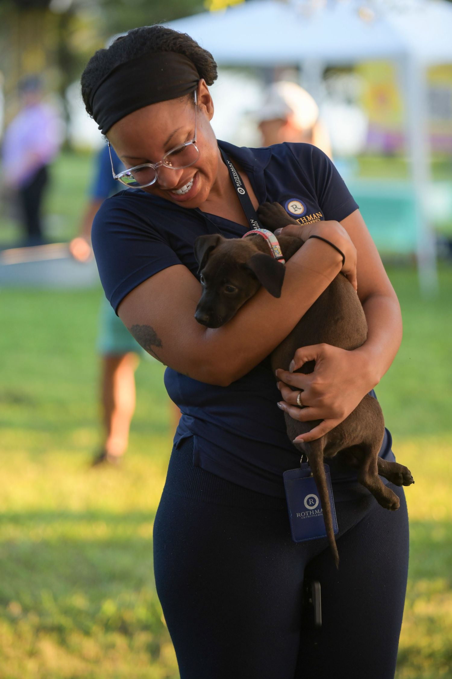 A smiling woman in navy blue hugs an adorable brown puppy outdoors at a sunny park event.