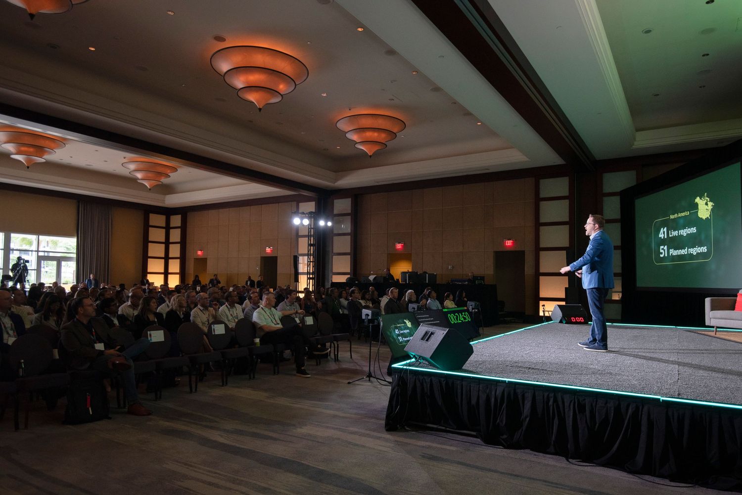 Speaker presents on stage at a corporate conference event with a large audience seated in a ballroom.