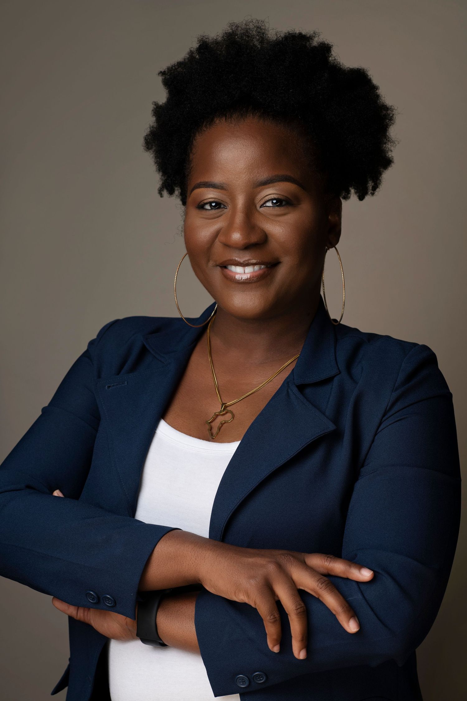 Professional Black woman in navy blazer smiling with arms crossed, natural hair, gold jewelry, studio portrait.